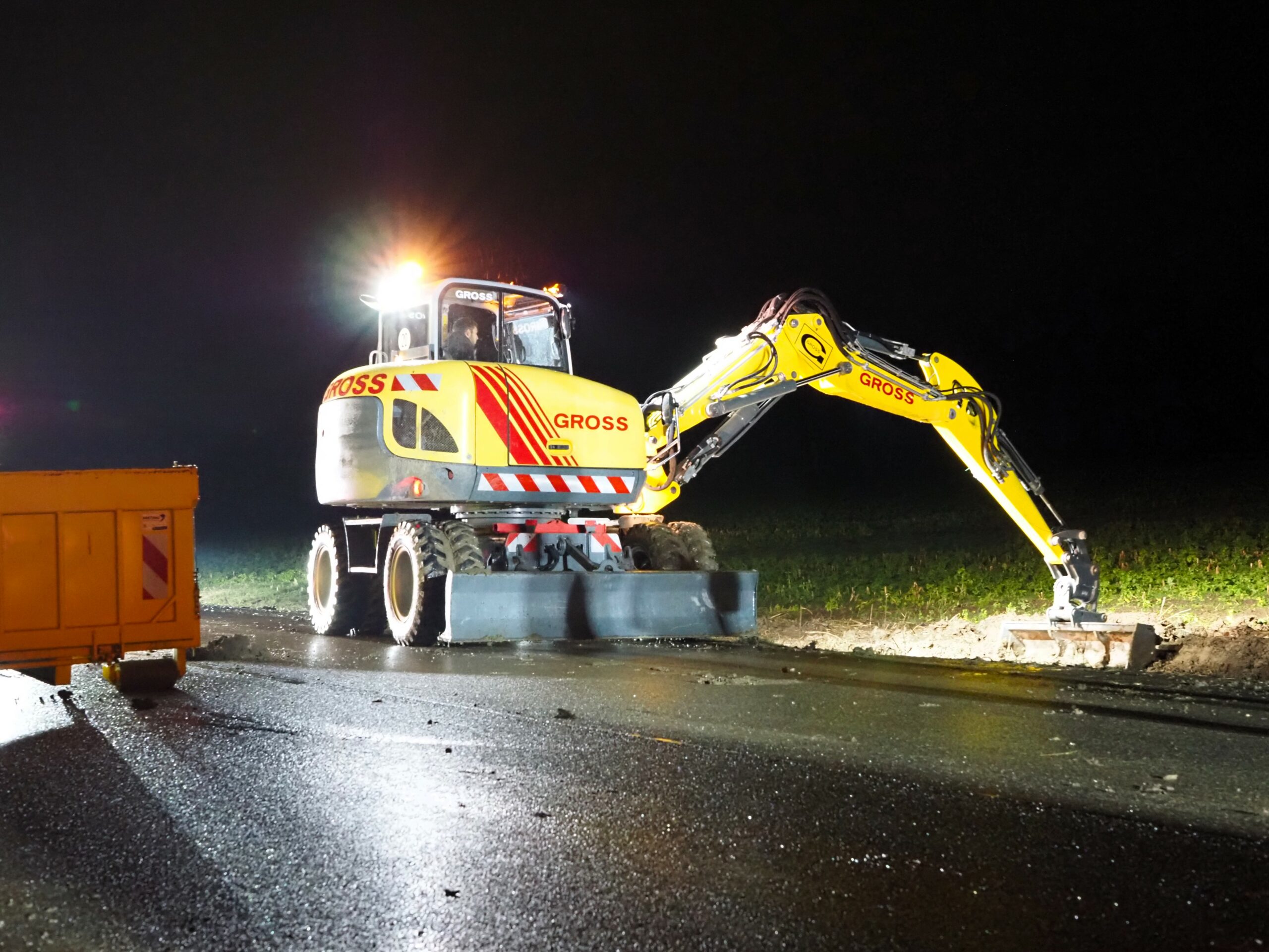 Nächtliche Baustelle: Ein beleuchteter gelber Bagger arbeitet an einer Straßenerneuerung.