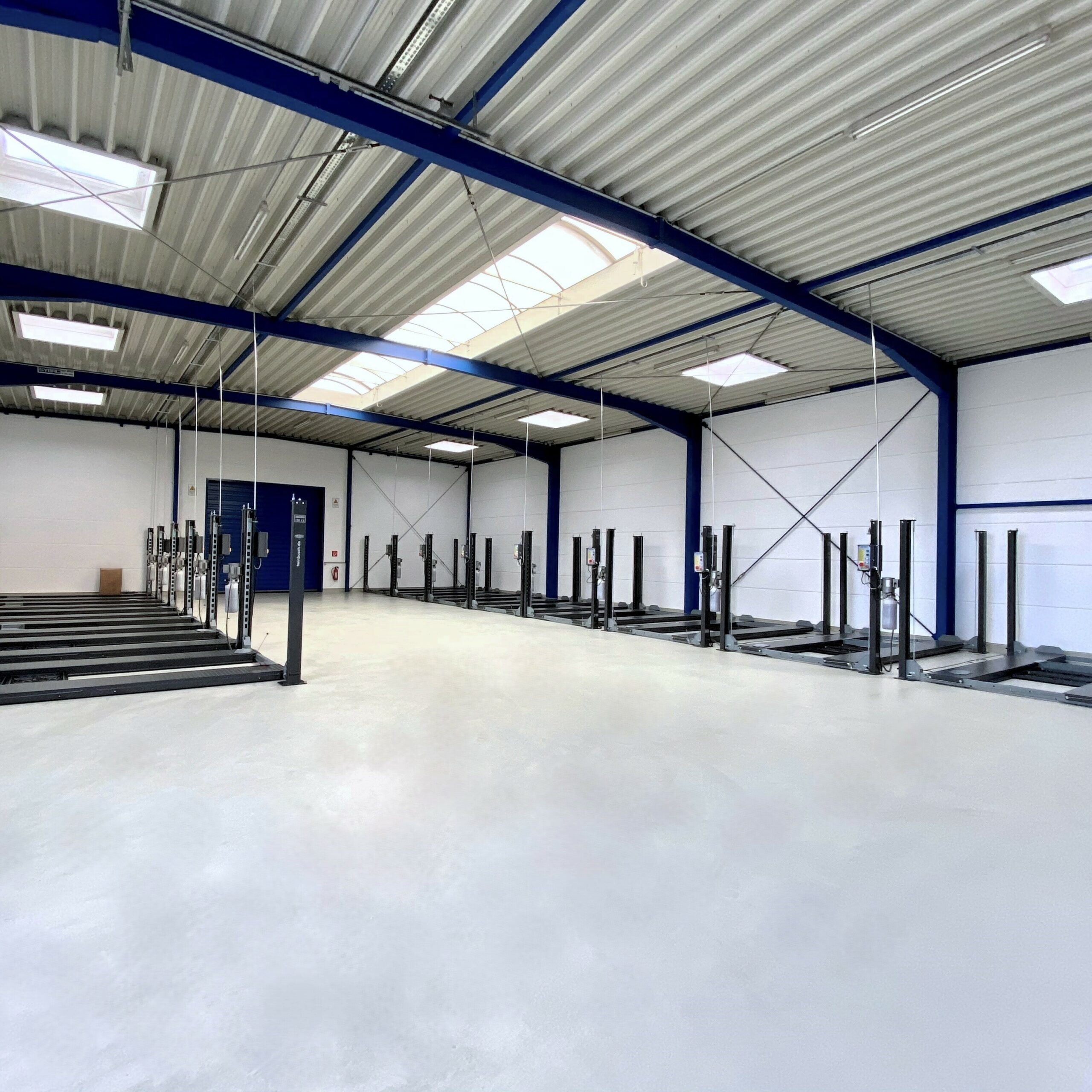 Große, leere Lagerhalle mit Stahlträgern, mehreren Autolifts und Oberlichtern; Wände und Decke sind weiß und blau.