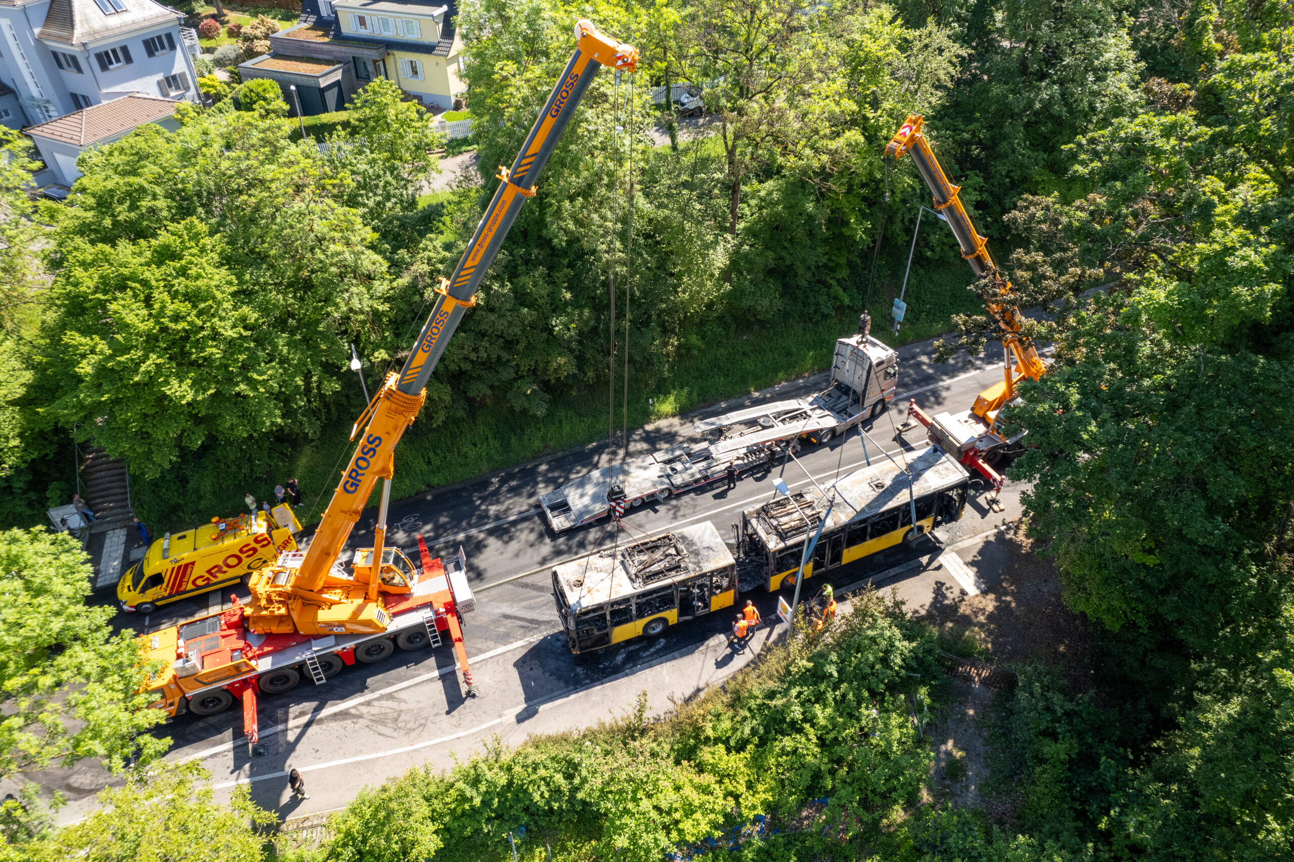 Zwei Kräne heben einen ausgebrannten Bus von der Straße, umgeben von Bäumen und Einsatzfahrzeugen.
