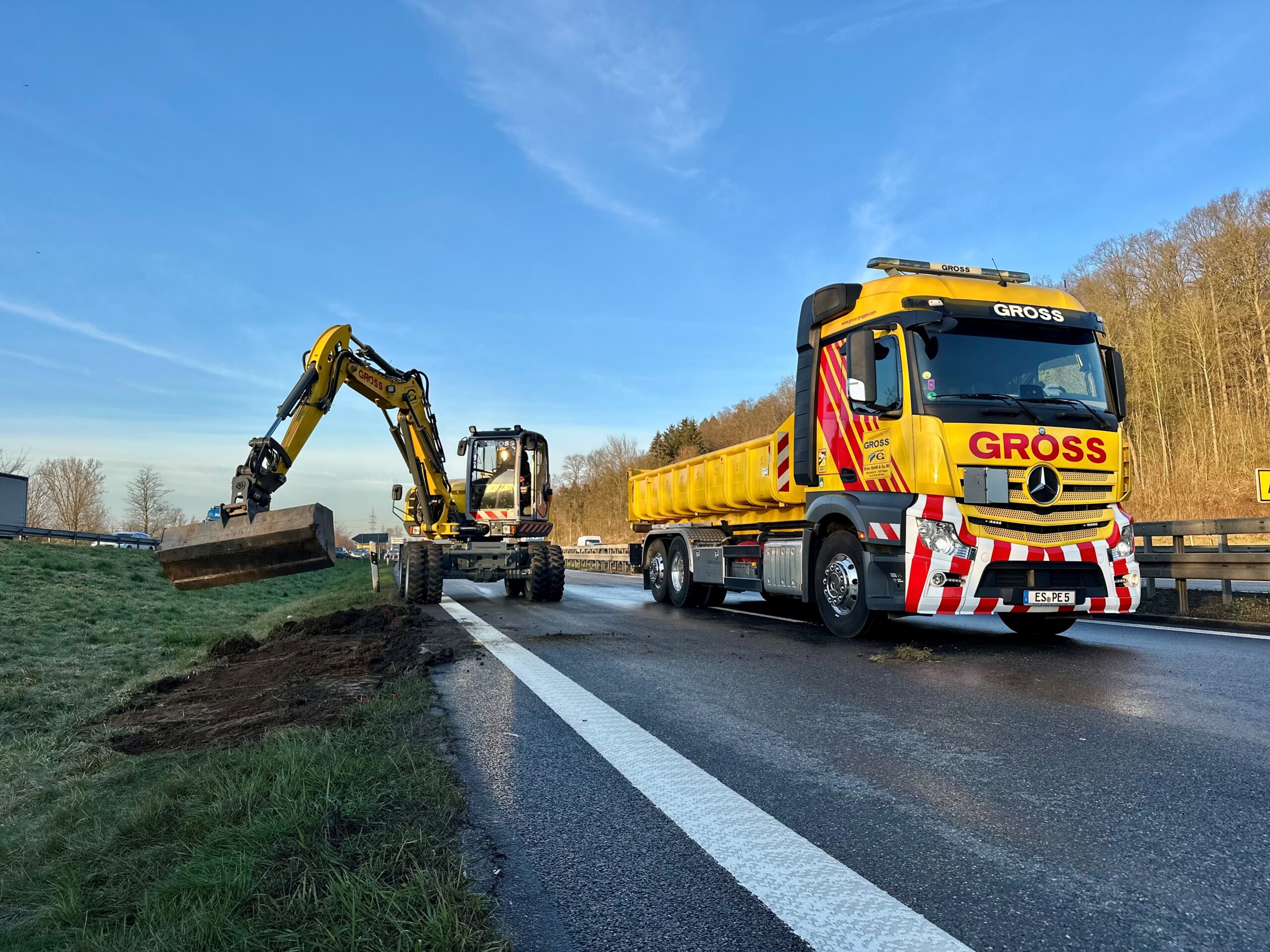 Gelber LKW und Bagger arbeiten auf einer Autobahn, graben Erde am Fahrbahnrand aus, blauer Himmel im Hintergrund.