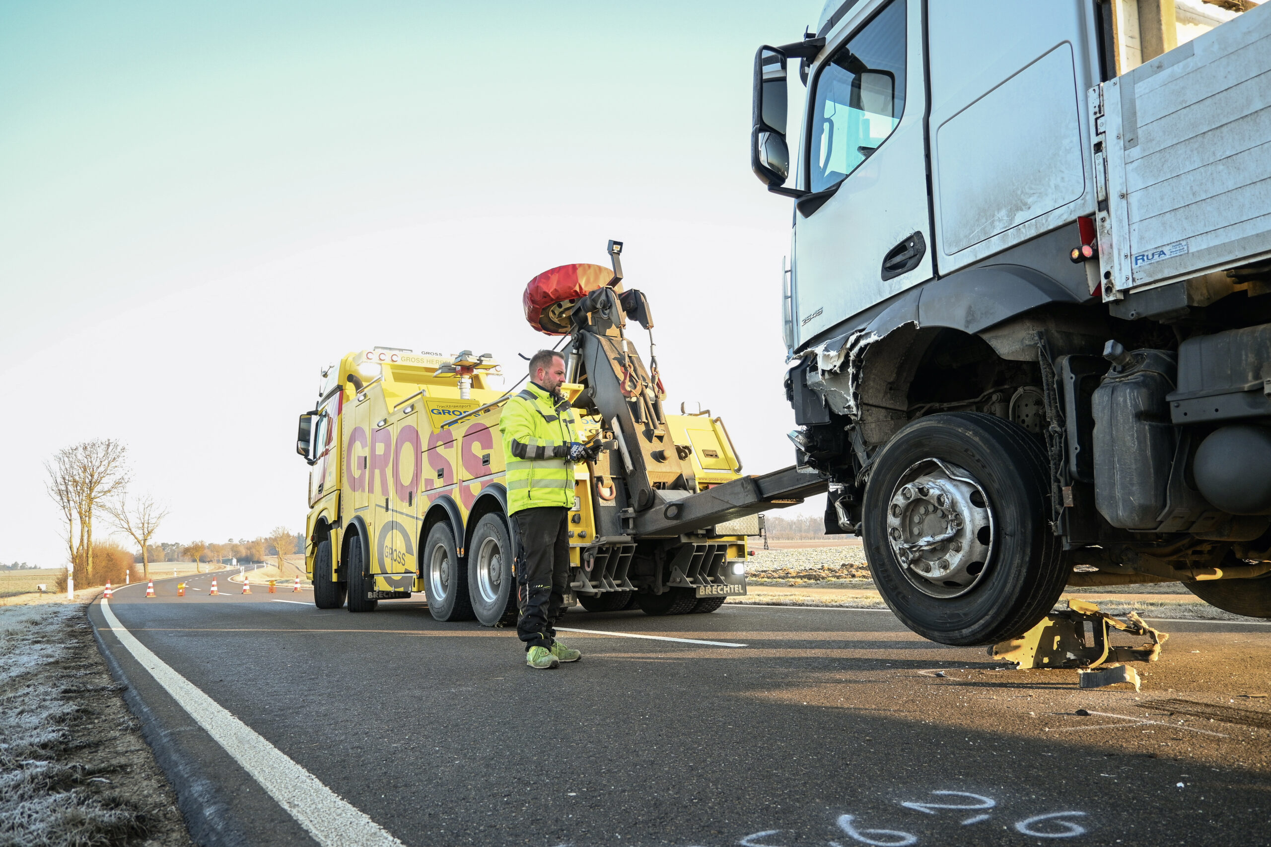 Abschleppwagen mit gelbem LKW hebt beschädigten weißen LKW auf Landstraße an, Arbeiter mit Warnweste vor Ort.