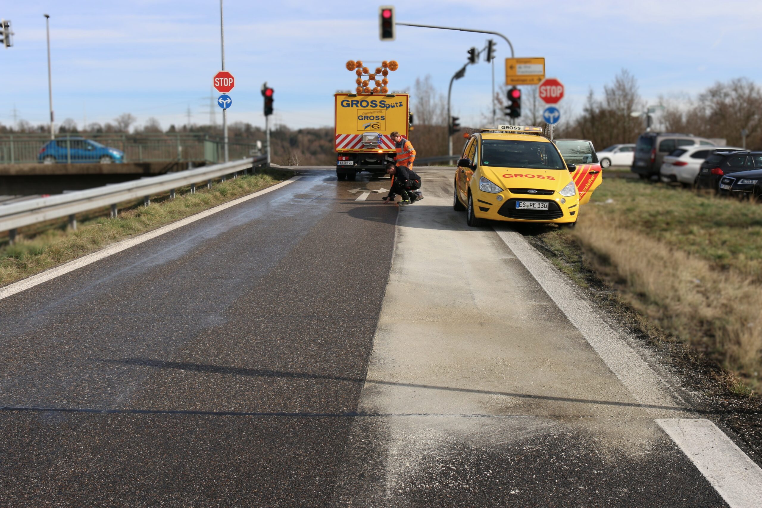 Straßenbaustelle mit zwei gelben Servicefahrzeugen. Arbeiter inspizieren die Fahrbahn, Stoppschild und Ampel im Hintergrund.