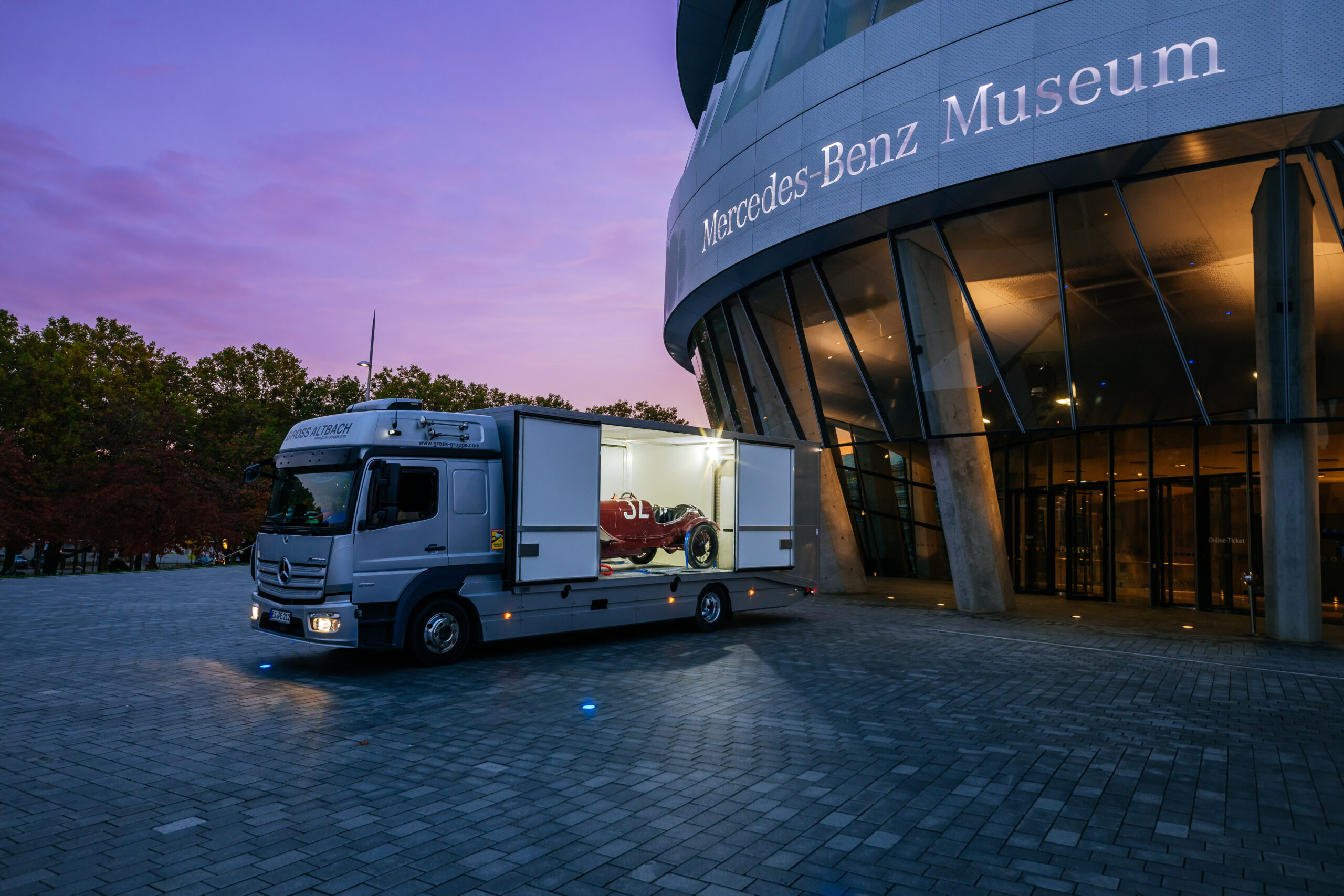 Lkw mit offenem Anhänger, der einen roten Rennwagen zeigt, vor dem Mercedes-Benz Museum bei Sonnenuntergang geparkt.