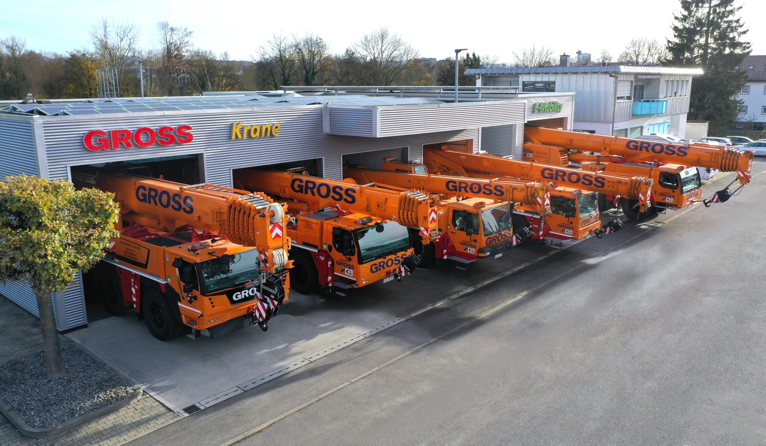 Sechs orange Kräne mit GROSS-Logo parken nebeneinander vor einem Gebäude mit der Aufschrift GROSS Krane.