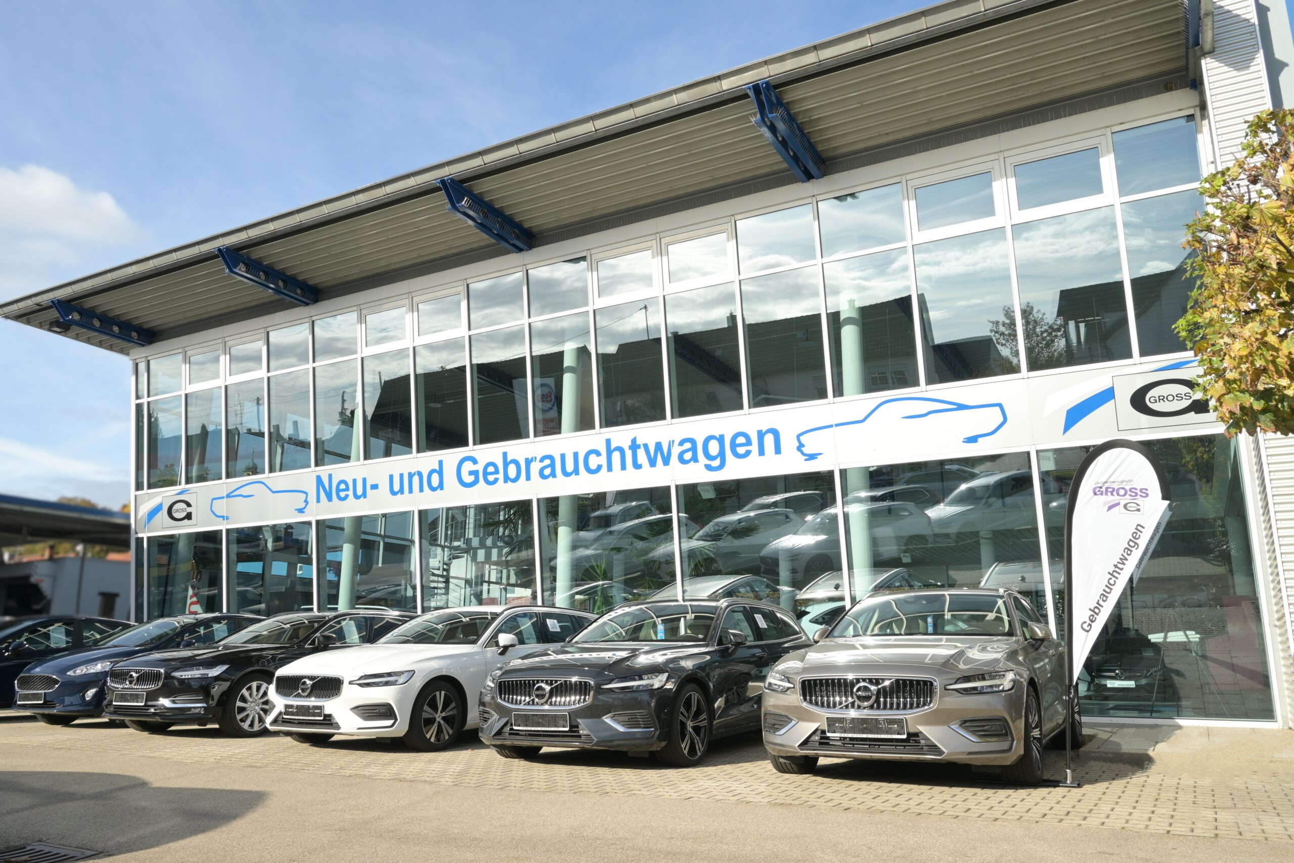 Autohaus mit Glasfassade, sechs geparkte Autos und einem Banner mit Gebrauchtwagen-Aufschrift im Sonnenschein.