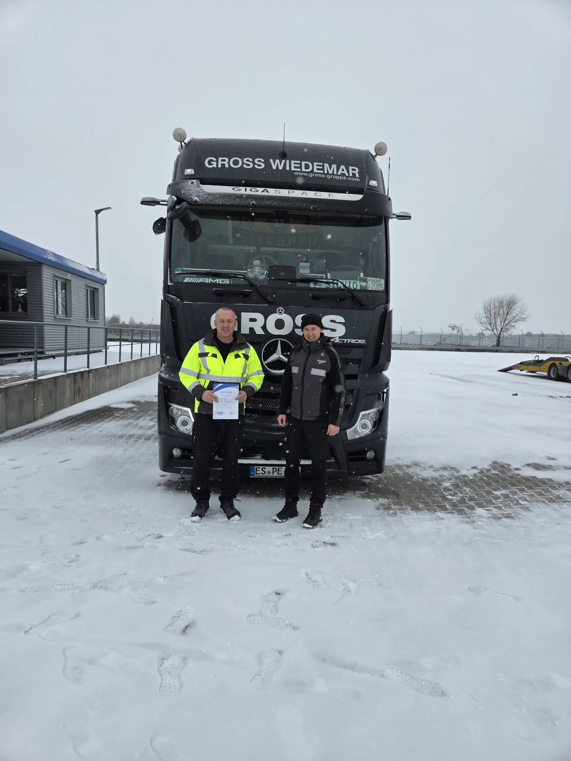 Zwei Personen stehen in Schneelandschaft vor einem großen Lkw mit der Aufschrift GROSS WIEDEMAR.