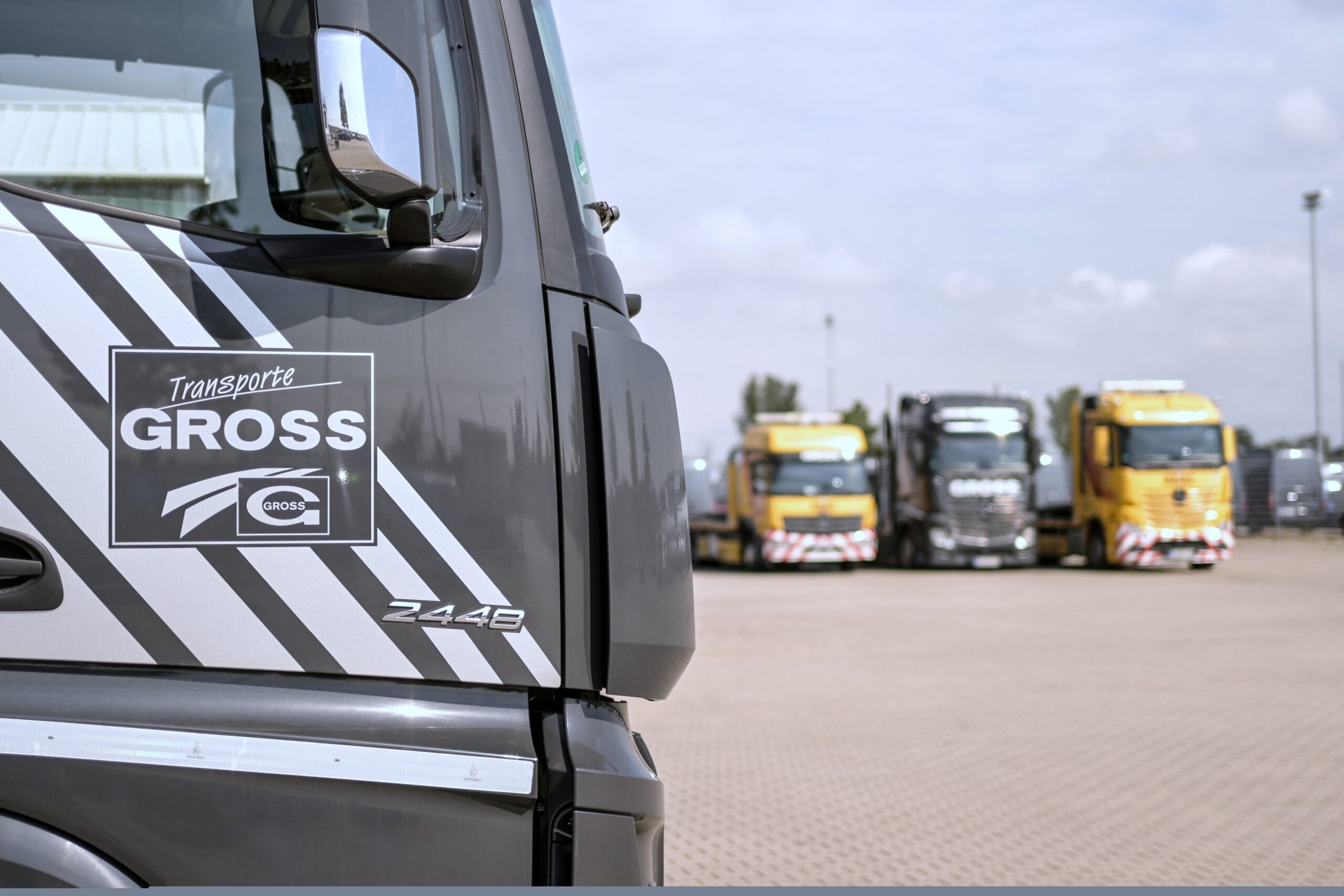 Nahaufnahme eines grauen Lastwagens mit GROSS-Logo, im Hintergrund drei unscharfe Lastwagen auf einem Parkplatz.