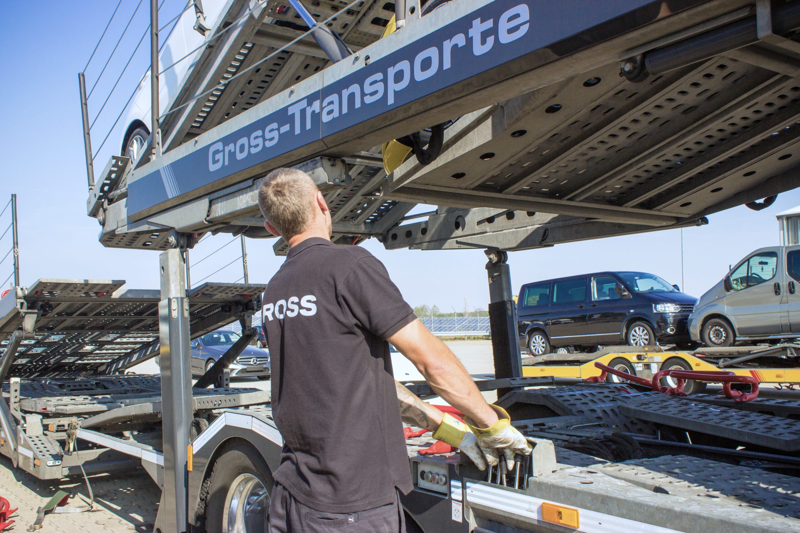 Ein Mann arbeitet an einem Autotransporter, trägt Handschuhe und schwarzes T-Shirt mit „GROSS“-Aufdruck, im Hintergrund Autos.