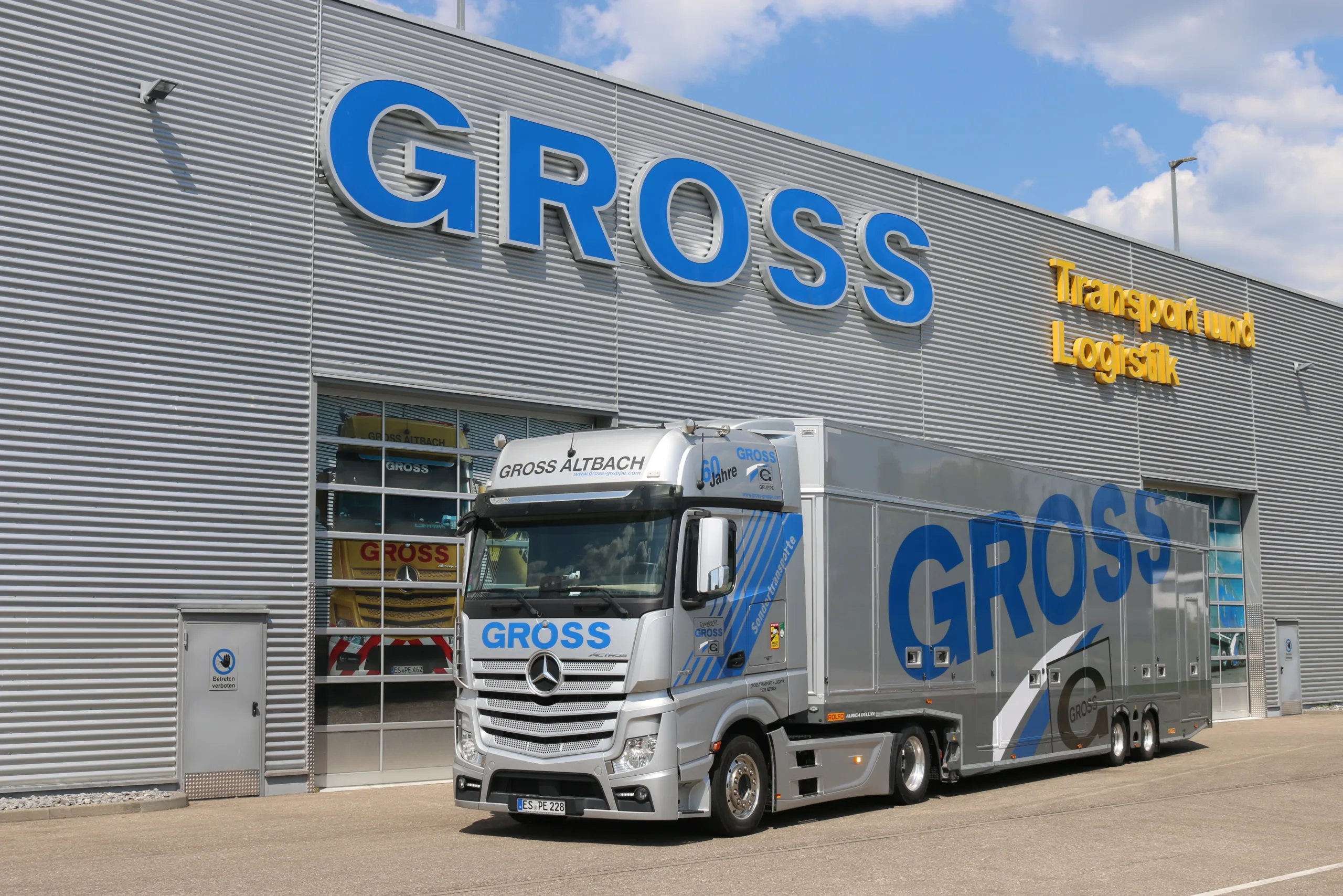 Silberner Lkw mit GROSS-Logo vor einem Gebäude mit der Aufschrift Transport und Logistik und klarem Himmel.