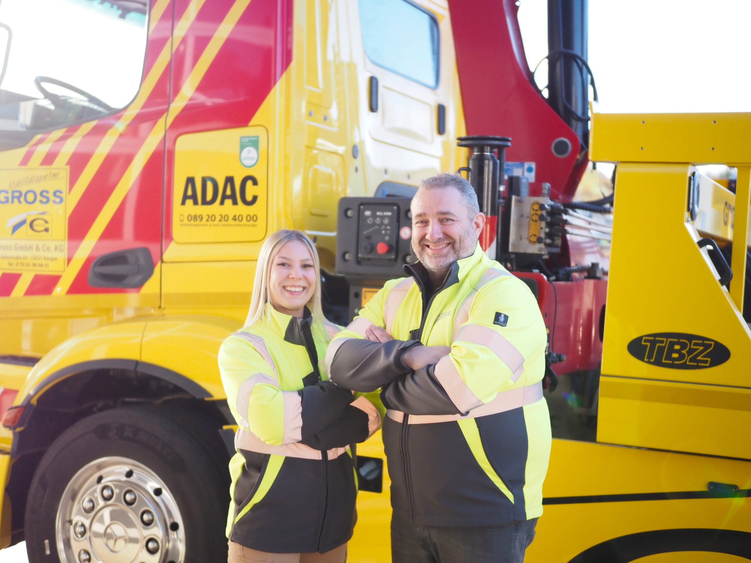 Zwei Personen in Warnjacken lächeln vor einem gelben Abschleppfahrzeug mit ADAC-Logo.