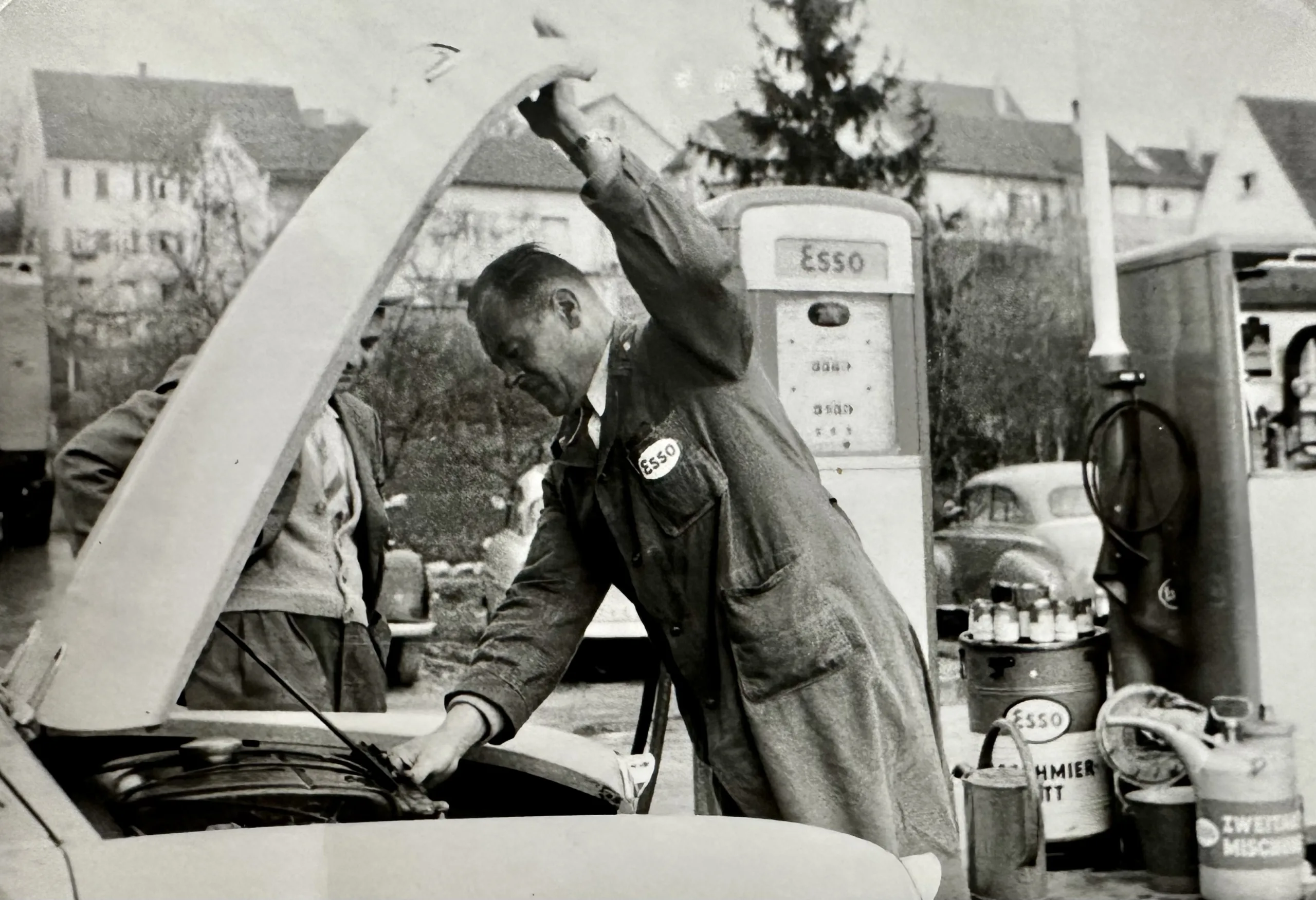 Mechaniker öffnet Motorhaube eines Oldtimers an einer Esso-Tankstelle in historischer Schwarz-Weiß-Aufnahme.