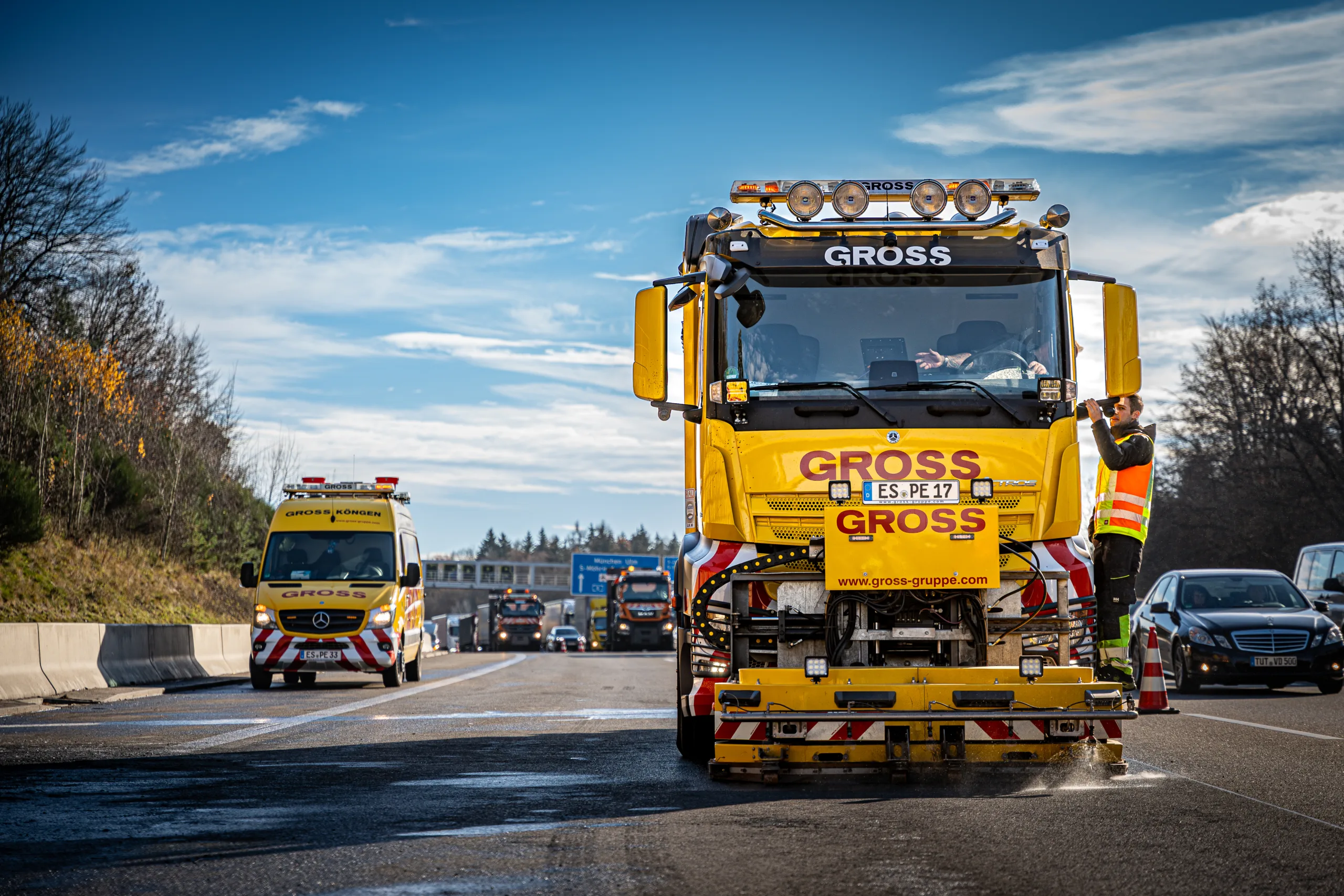 Gelbes Straßenreinigungsfahrzeug mit Arbeiter in Warnweste, begleitet von einem Rettungsfahrzeug auf einer Autobahn.