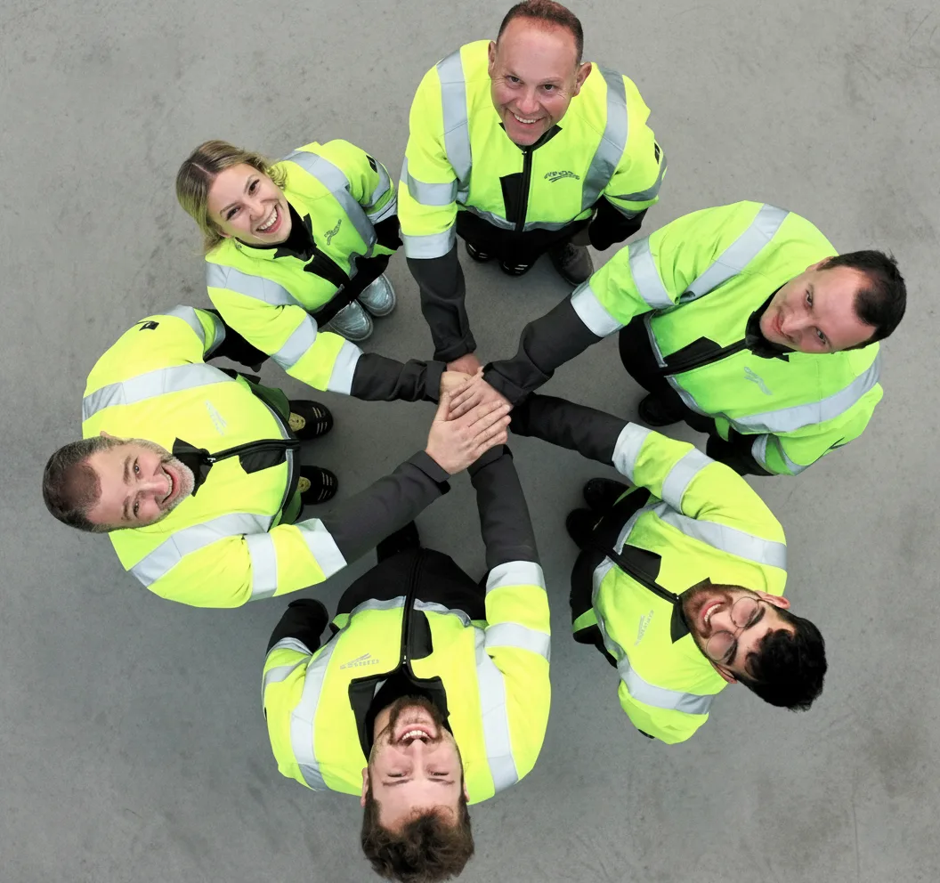 Sechs Personen in leuchtend gelben Jacken bilden einen Kreis und legen die Hände übereinander, lächelnd.