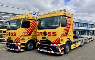 Zwei gelbe Lkw mit ADAC-Logos stehen vor einem modernen Logistikgebäude, umgeben von Wolken.