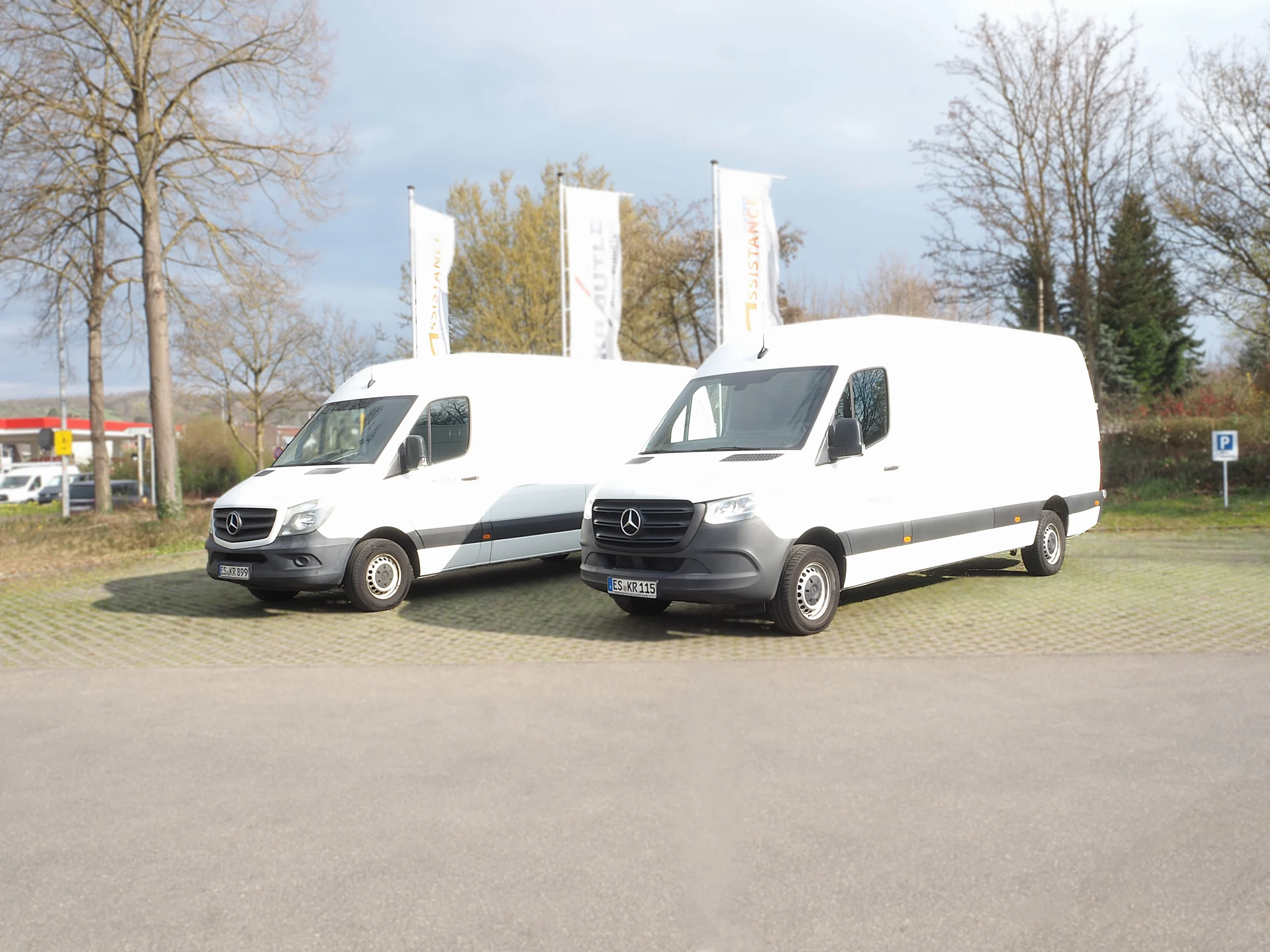 Zwei weiße Mercedes-Vans stehen auf einem Parkplatz, umgeben von Bäumen und Werbefahnen.