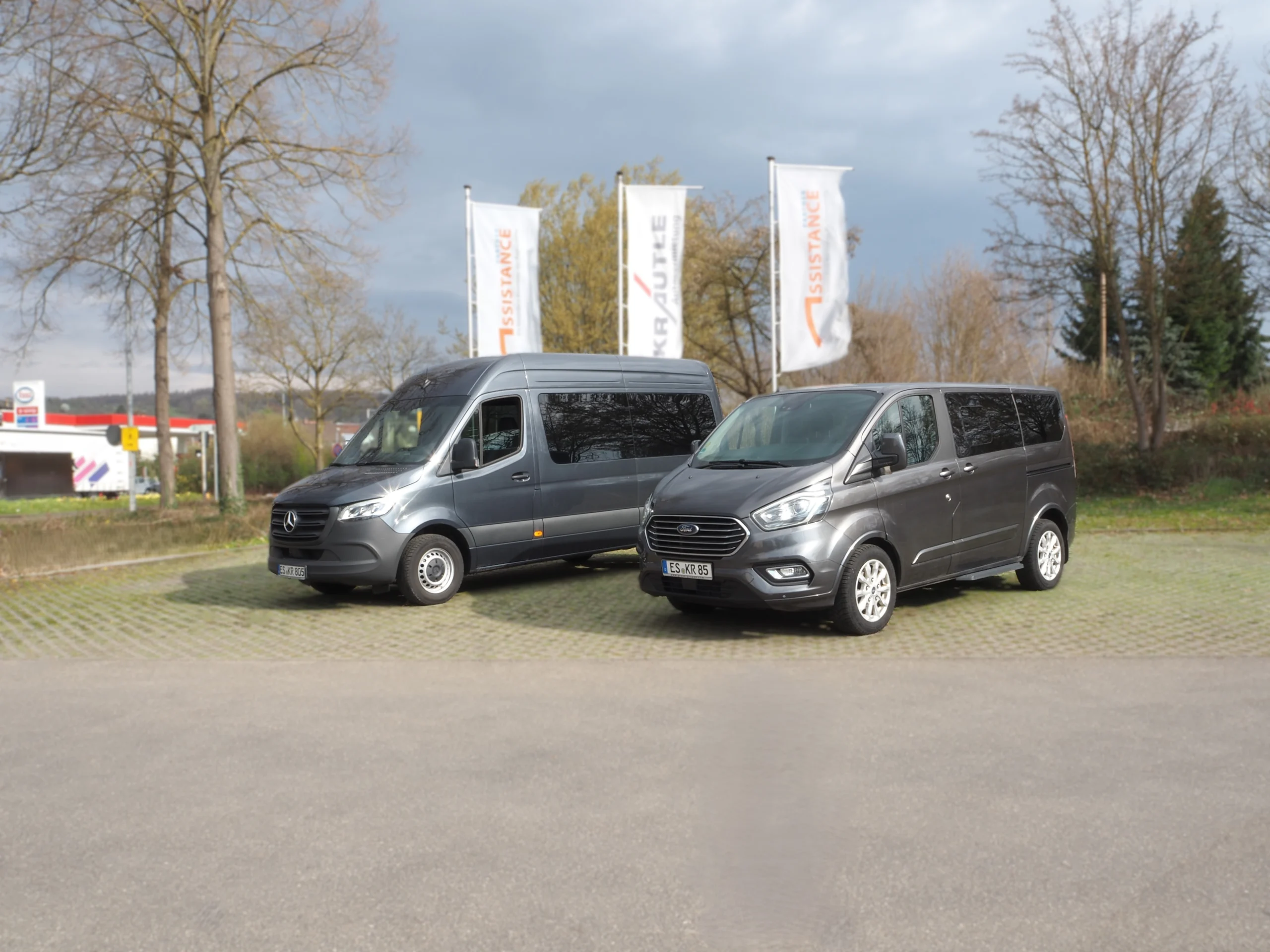 Zwei graue Nutzfahrzeuge, ein Mercedes Sprinter und ein Ford Tourneo, stehen auf einem gepflasterten Parkplatz.