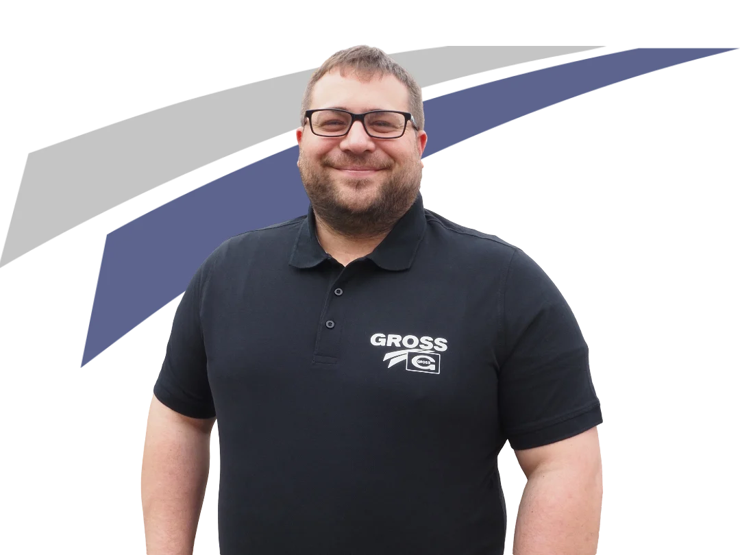 Ein lächelnder Mann mit Brille trägt ein schwarzes Polo-Shirt mit dem Aufdruck „GROSS“ vor einem grafischen Hintergrund.