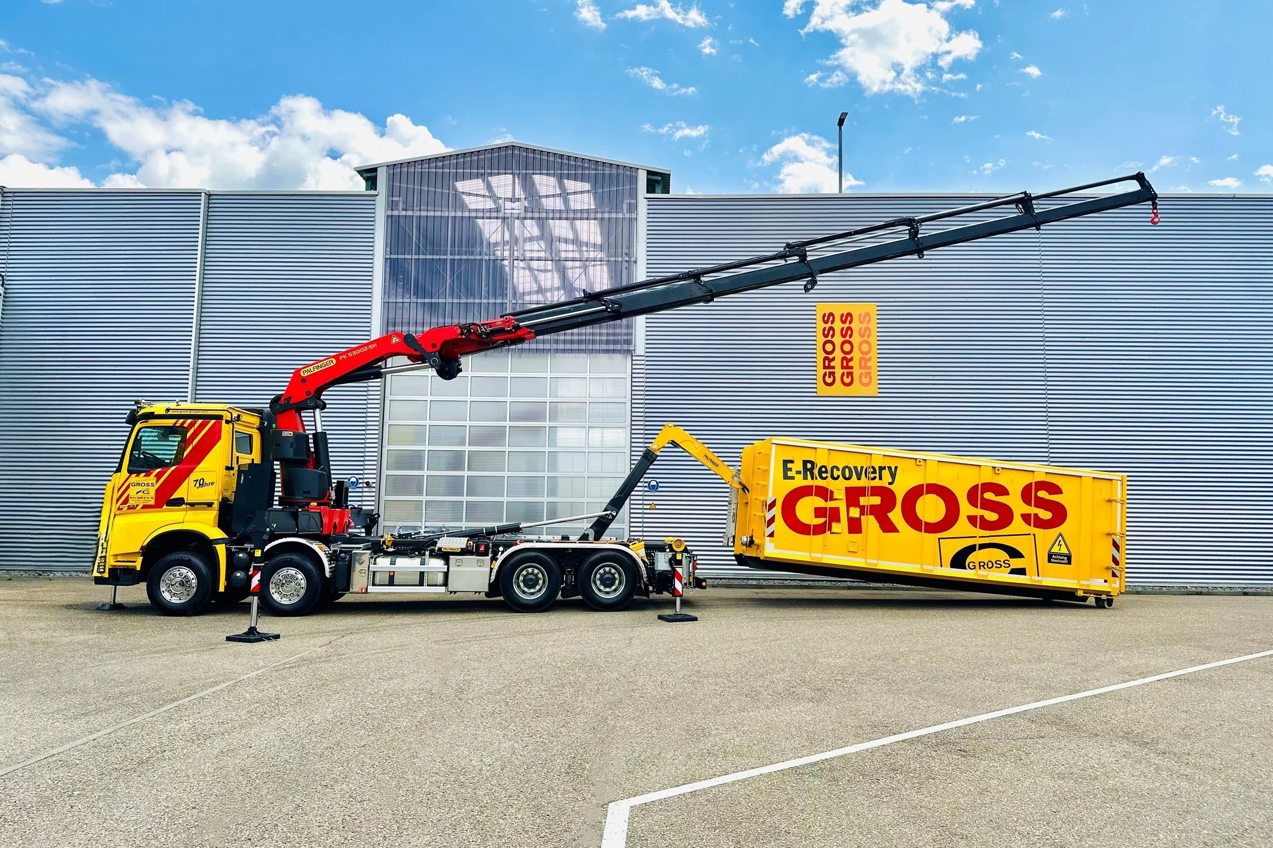 Ein gelber Lkw mit Kran hebt einen großen Container vor einem modernen Gebäude. Auf dem Container steht E-Recovery GROSS.
