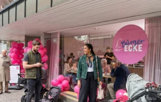 Junge Frau und Mann stehen vor einem Café mit pinken Luftballons. Im Hintergrund sitzen Gäste und genießen Kuchen.