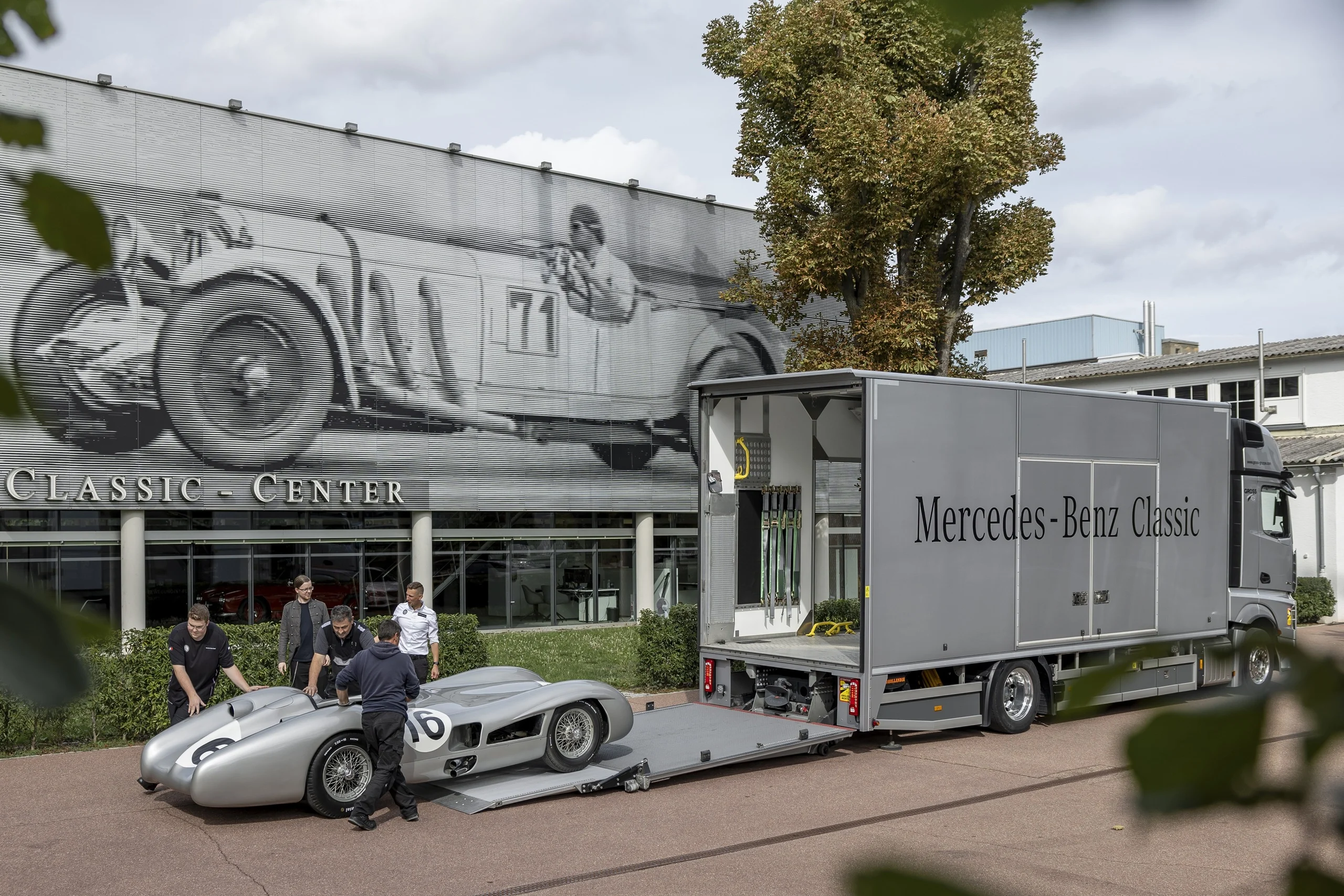 Zwei Personen laden einen silbernen Oldtimer vor dem Mercedes-Benz Classic Center auf einen Transporter.