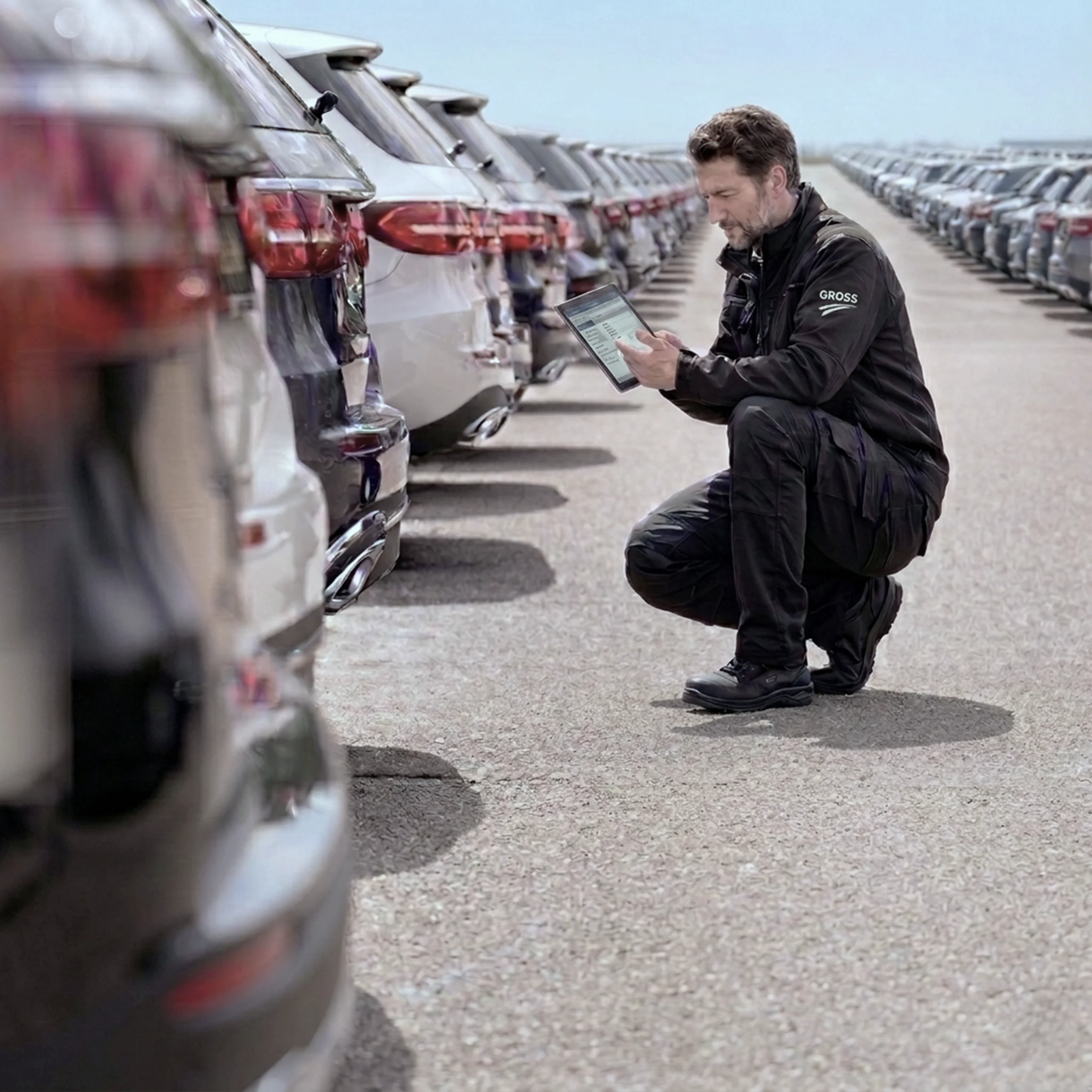Ein Mann in Arbeitskleidung kniet vor mehreren Autos und nutzt ein Tablet zur Fahrzeugüberprüfung auf einem Parkplatz.