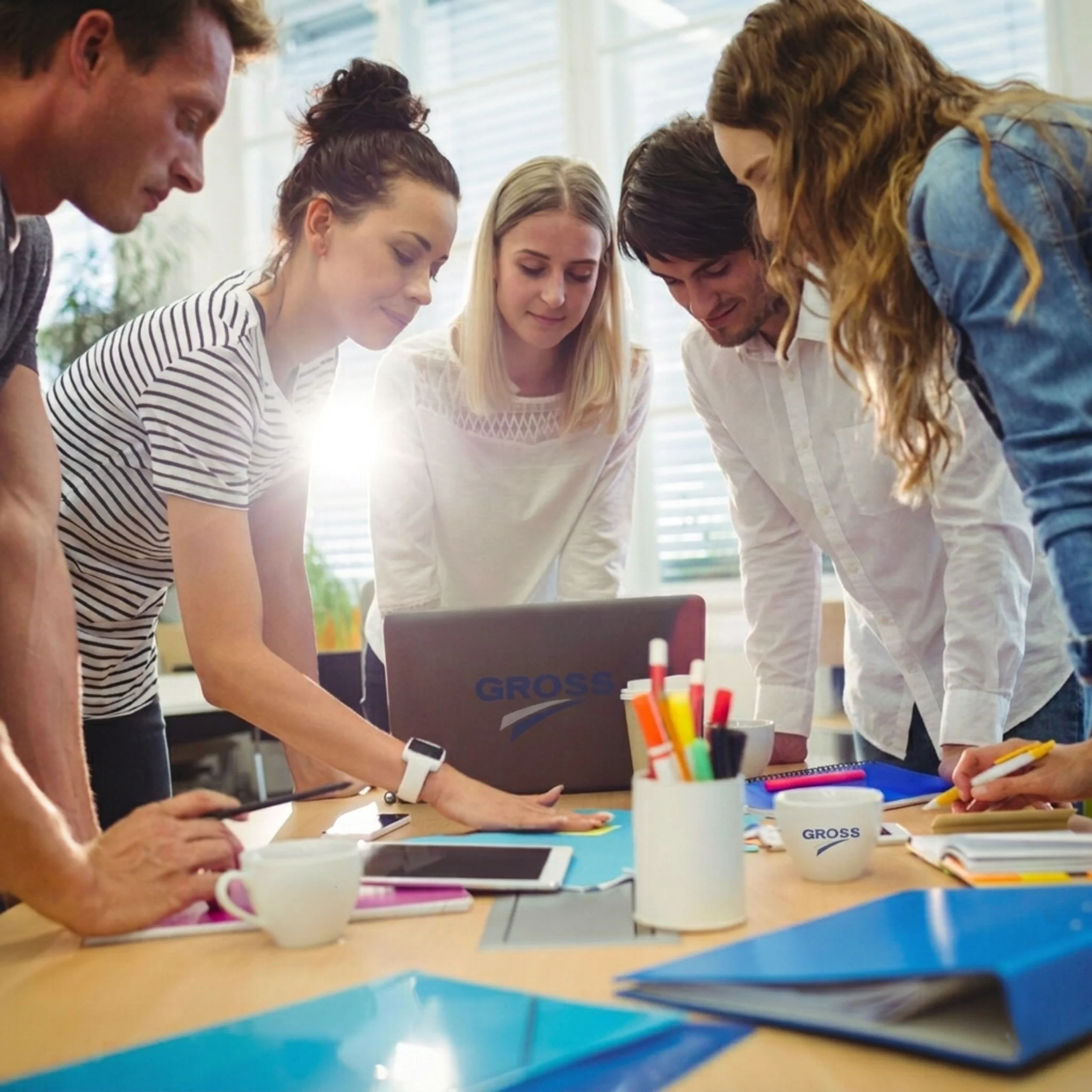 Fünf Personen arbeiten an einem Tisch, schauen auf ein Laptop und besprechen Ideen, umgeben von Bürobedarf.
