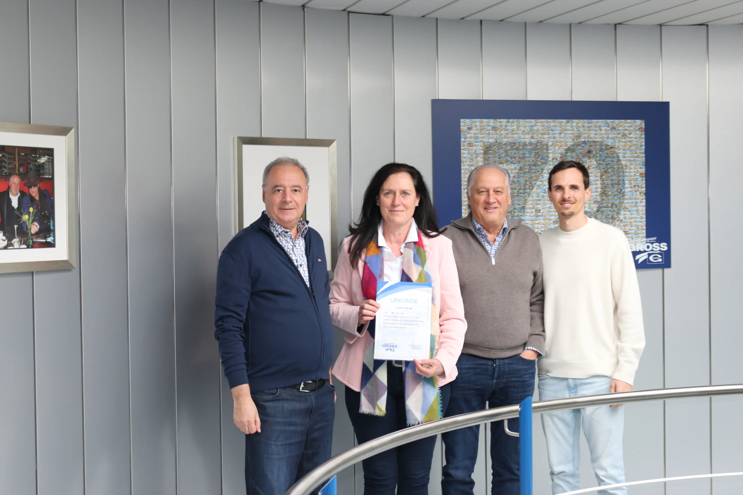 Drei Männer und eine Frau halten eine Urkunde in einem modernen Gebäude, umgeben von Bildern und dekorativen Wänden.
