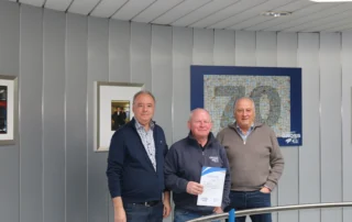 Drei Männer stehen vor einer Wand mit Bildern und einem blauen Kunstwerk, einer hält eine Urkunde in den Händen.