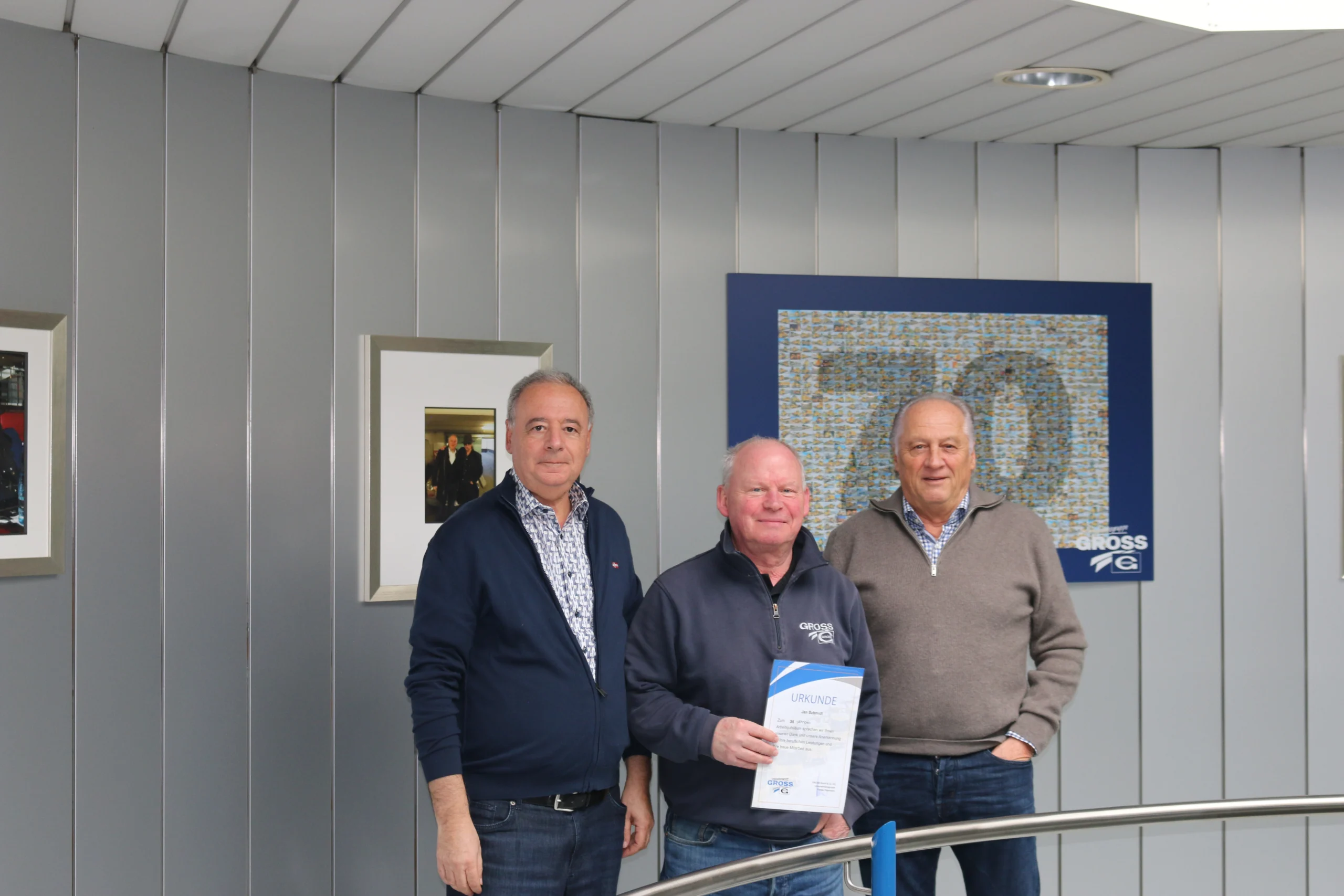 Drei Männer stehen vor einer Wand mit Bildern und einem blauen Kunstwerk, einer hält eine Urkunde in den Händen.
