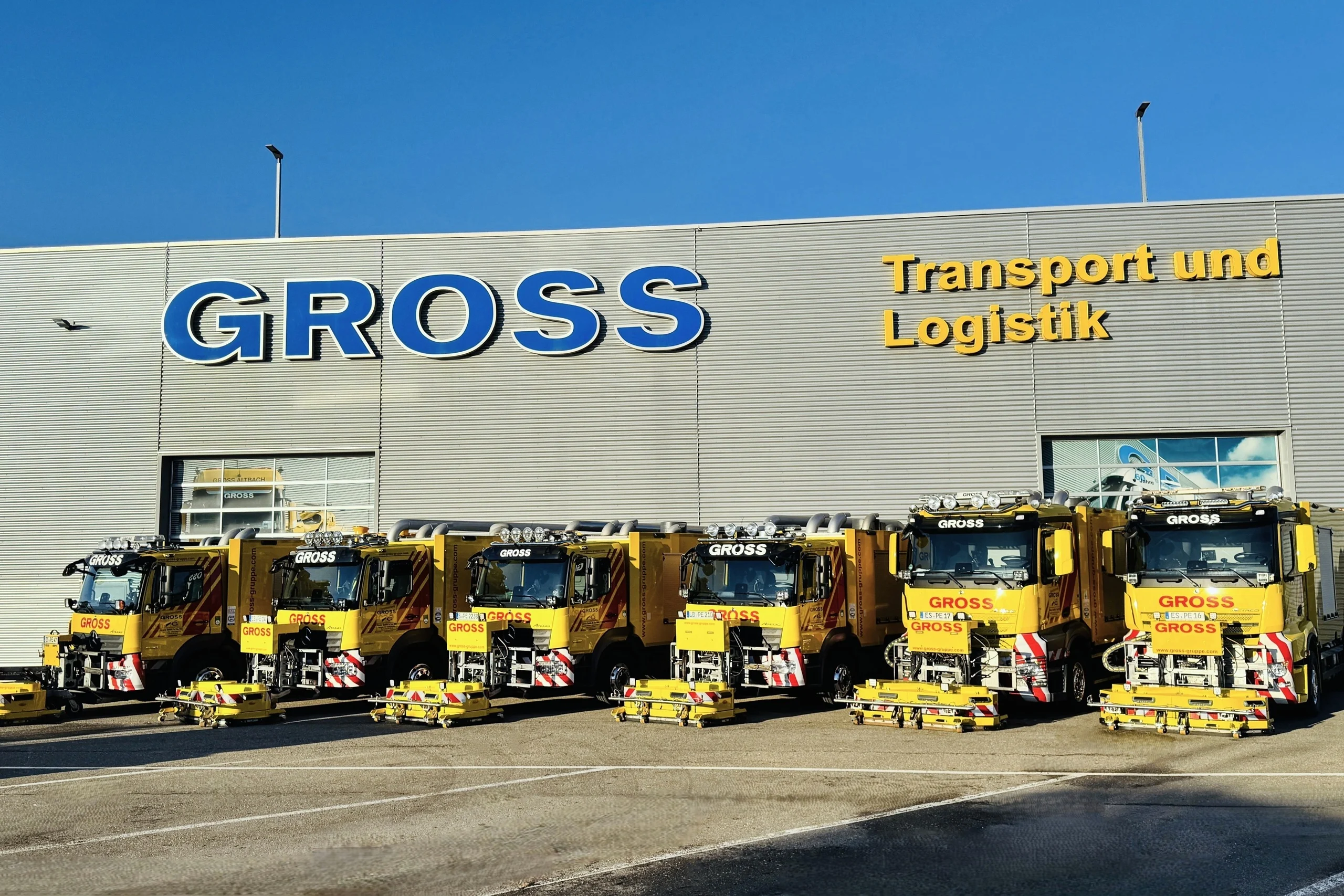 Fünf gelbe Lkw mit GROSS-Aufschrift stehen vor einem modernen Firmengebäude mit blauen und gelben Schriftzügen.