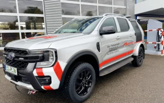 Grauer SUV mit roten und weißen Streifen, geparkt vor einem Gebäude. Schriftzüge auf der Seite und offroad-tauglichen Reifen.