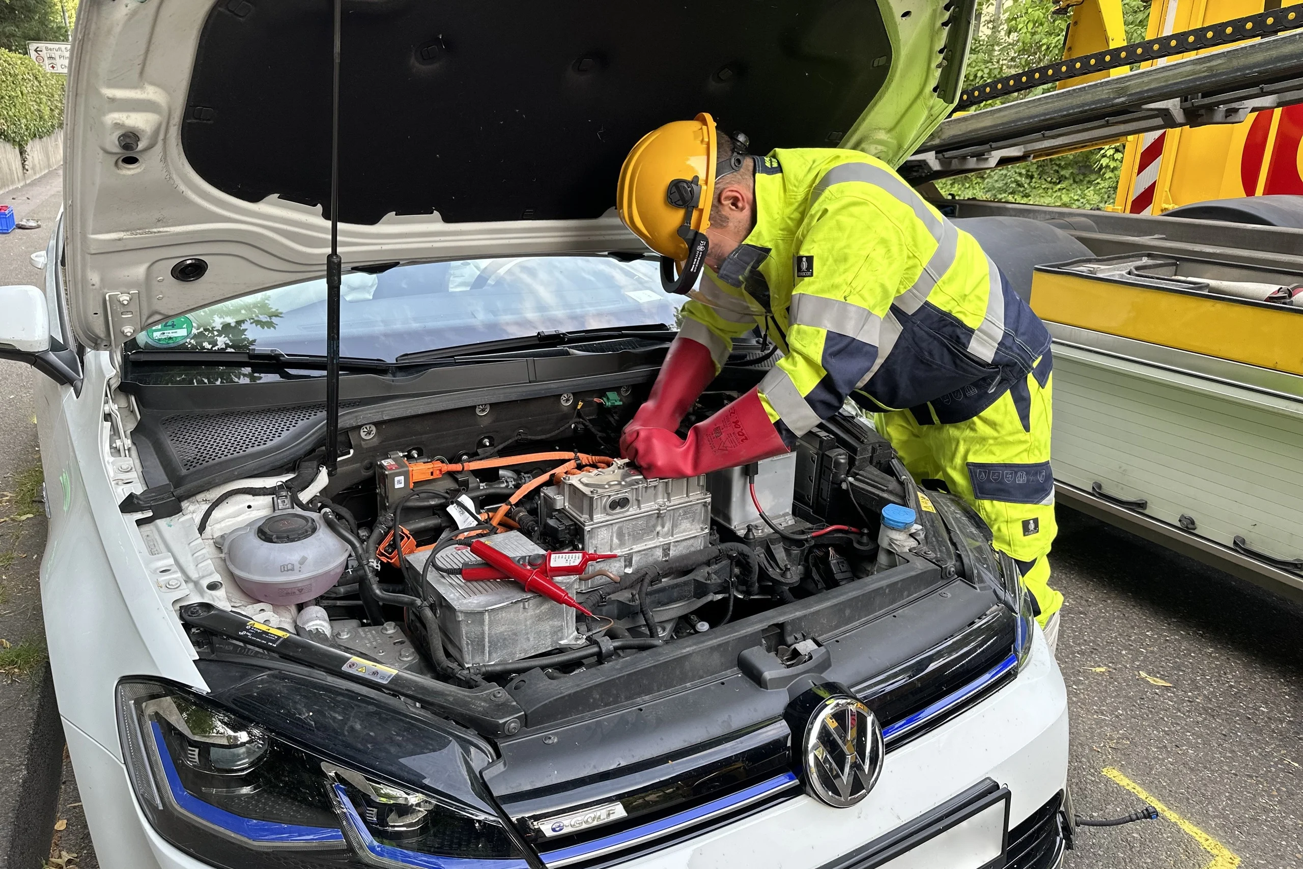 Mechaniker mit gelber Warnjacke und Helm repariert Elektrokomponenten im Motorraum eines weißen Volkswagen.