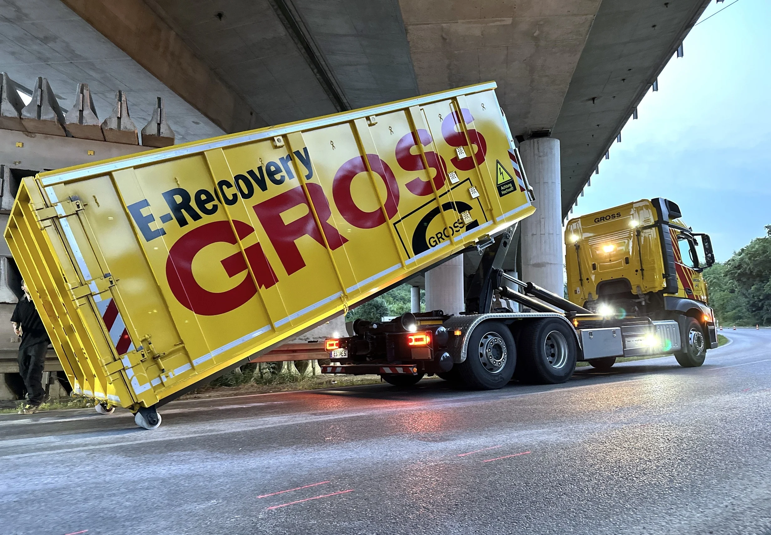 Gelber Lkw mit der Aufschrift E-Recovery GROSS hebt Abrollcontainer unter einer Brücke an, im Hintergrund Wald.