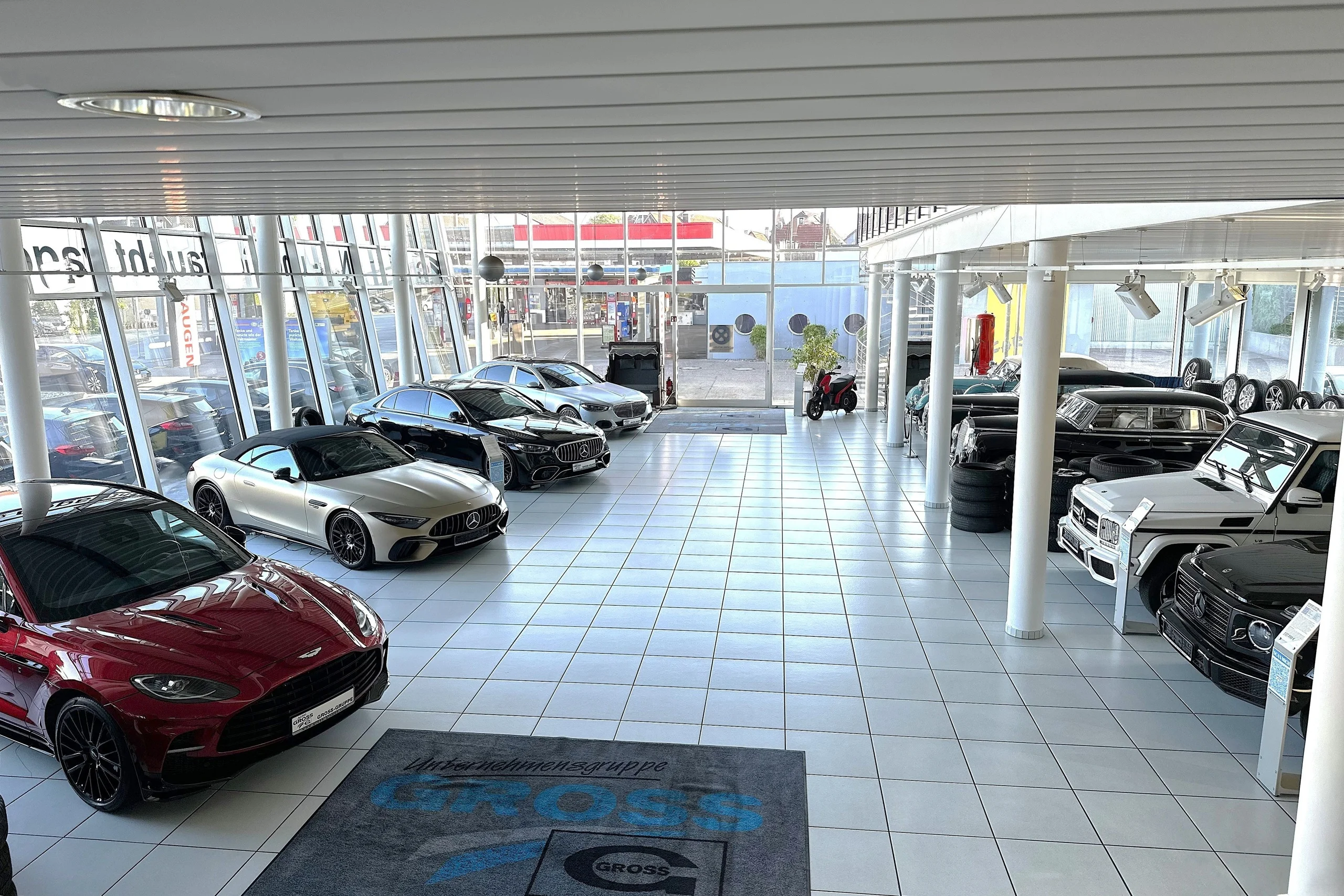 Fünf Luxusautos stehen in einem modernen Autohaus mit großen Fenstern und grauem Boden.