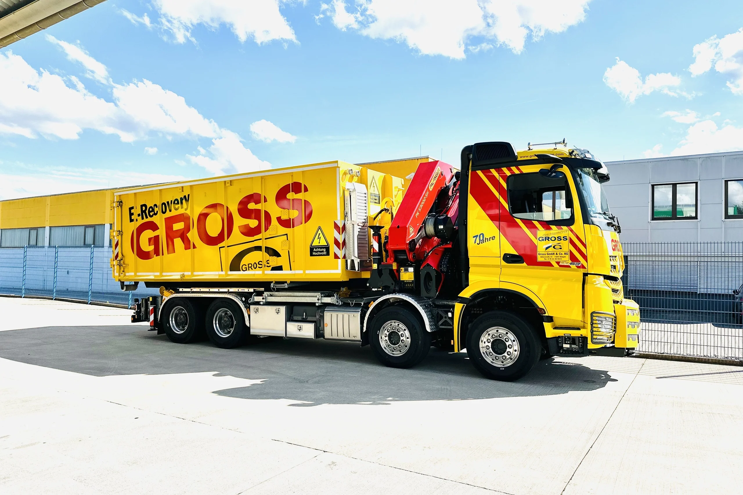 Gelber Lkw mit Montagekran und Aufschrift E-Recovery GROSS parkt vor einer Lagerhalle unter blauem Himmel.