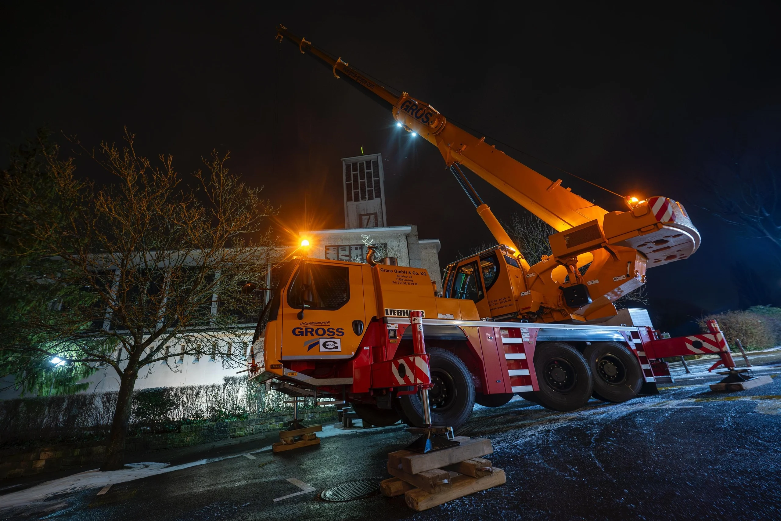 Kran in der Nacht: orangefarbenes Fahrzeug mit ausfahrbarem Arm steht vor einem Gebäude, um Bauarbeiten durchzuführen.