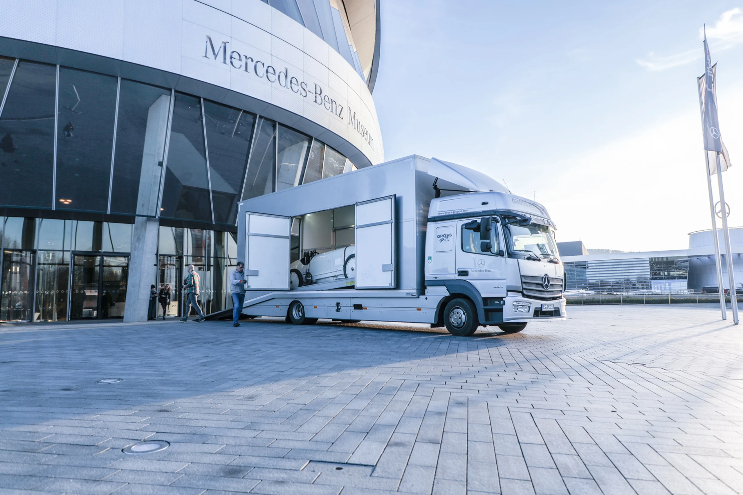 Lkw vor dem Mercedes-Benz Museum, bereit, ein Fahrzeug abzuladen. Menschen stehen in der Nähe.