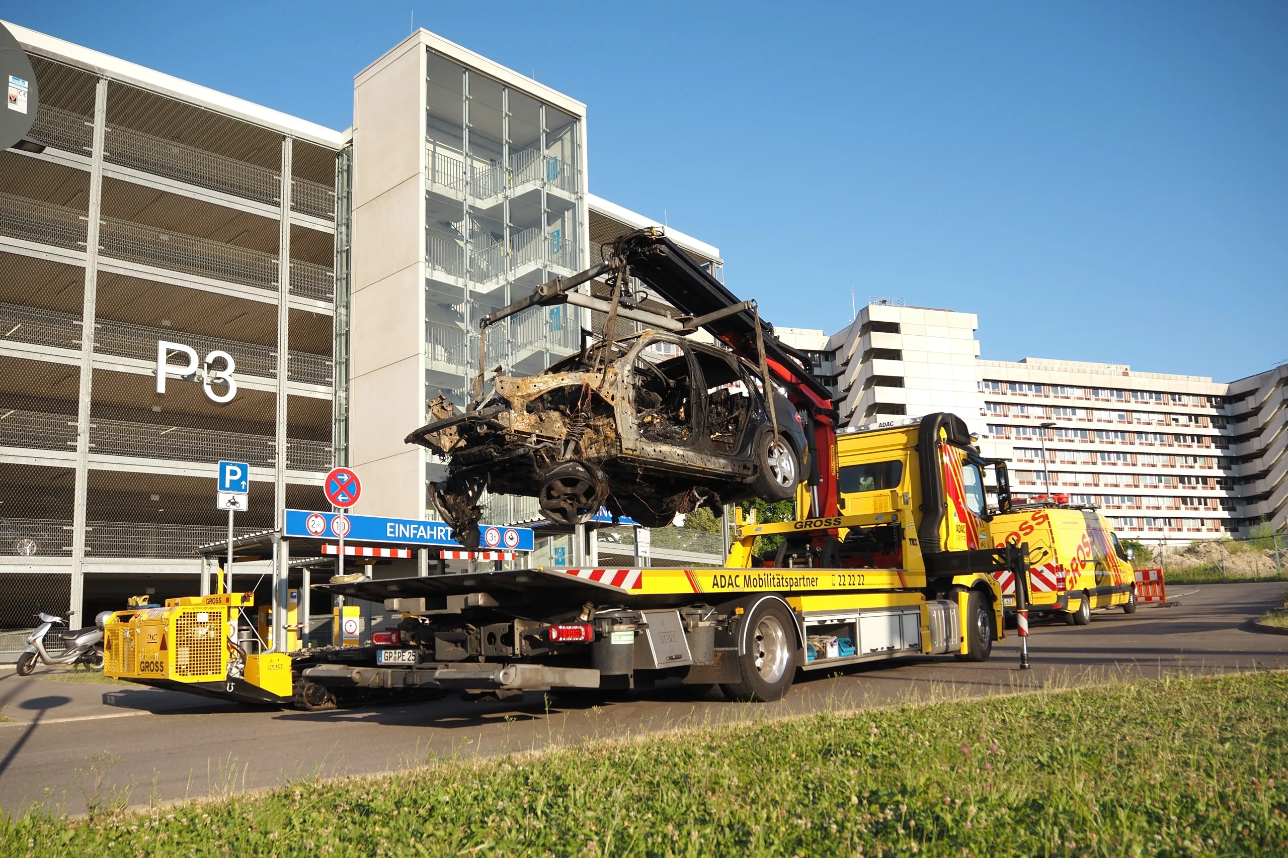 Abschleppwagen hebt ein ausgebranntes Auto neben einem Parkhaus und einem Wohnkomplex hoch.