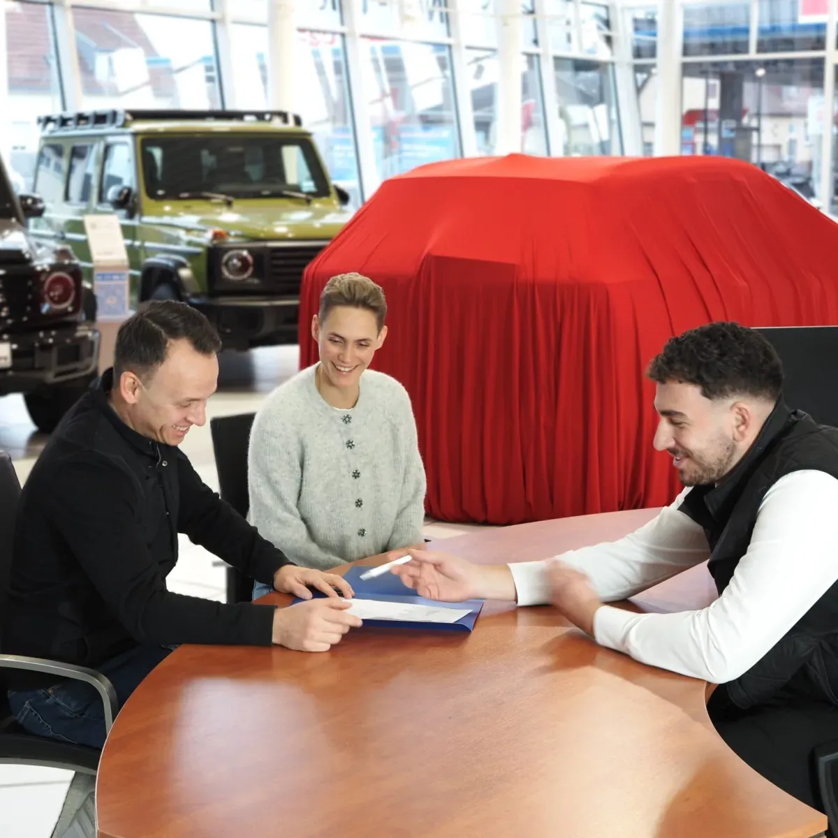 Drei Männer sitzen an einem Tisch in einem Autohaus, lachen und unterhalten sich über einen Vertrag, im Hintergrund Autos.