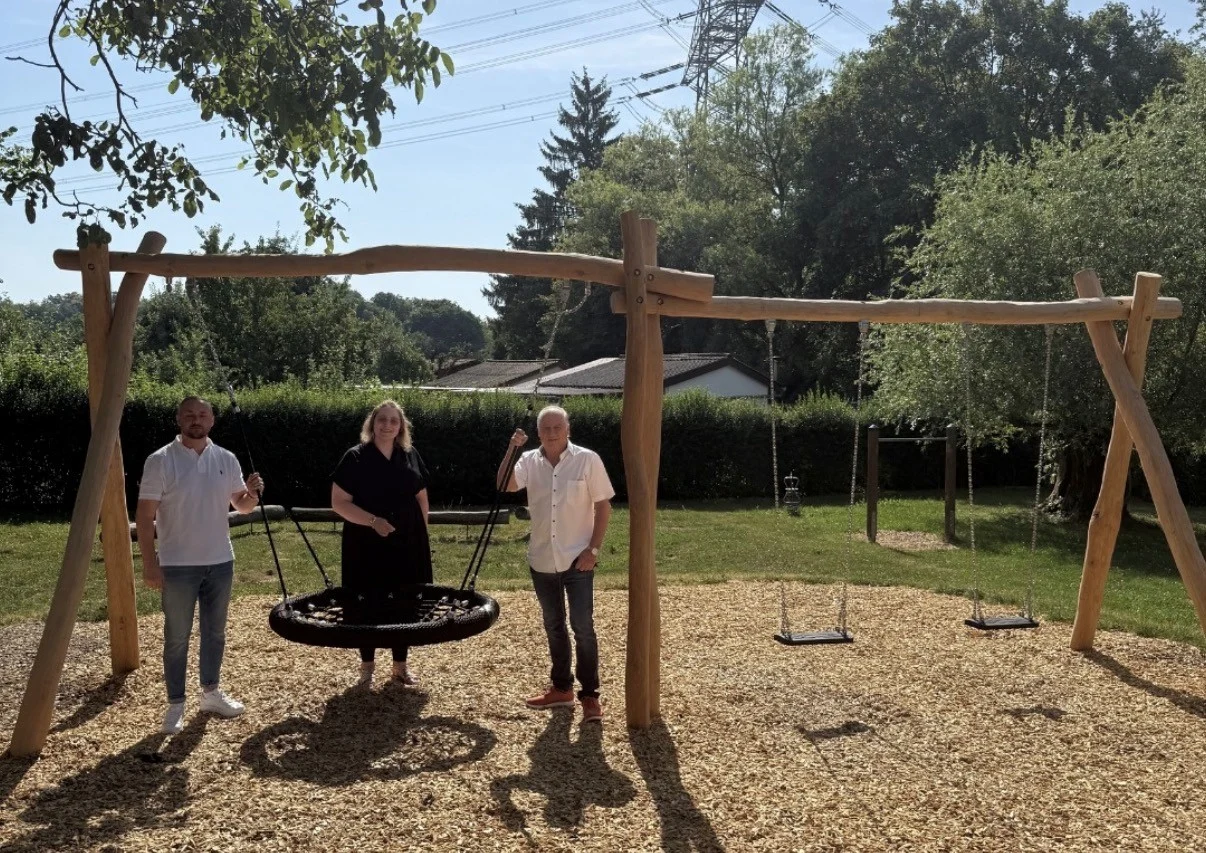 Drei Personen stehen auf einem Spielplatz neben einer großen, runden Schaukel und zwei kleinen Schwingen.
