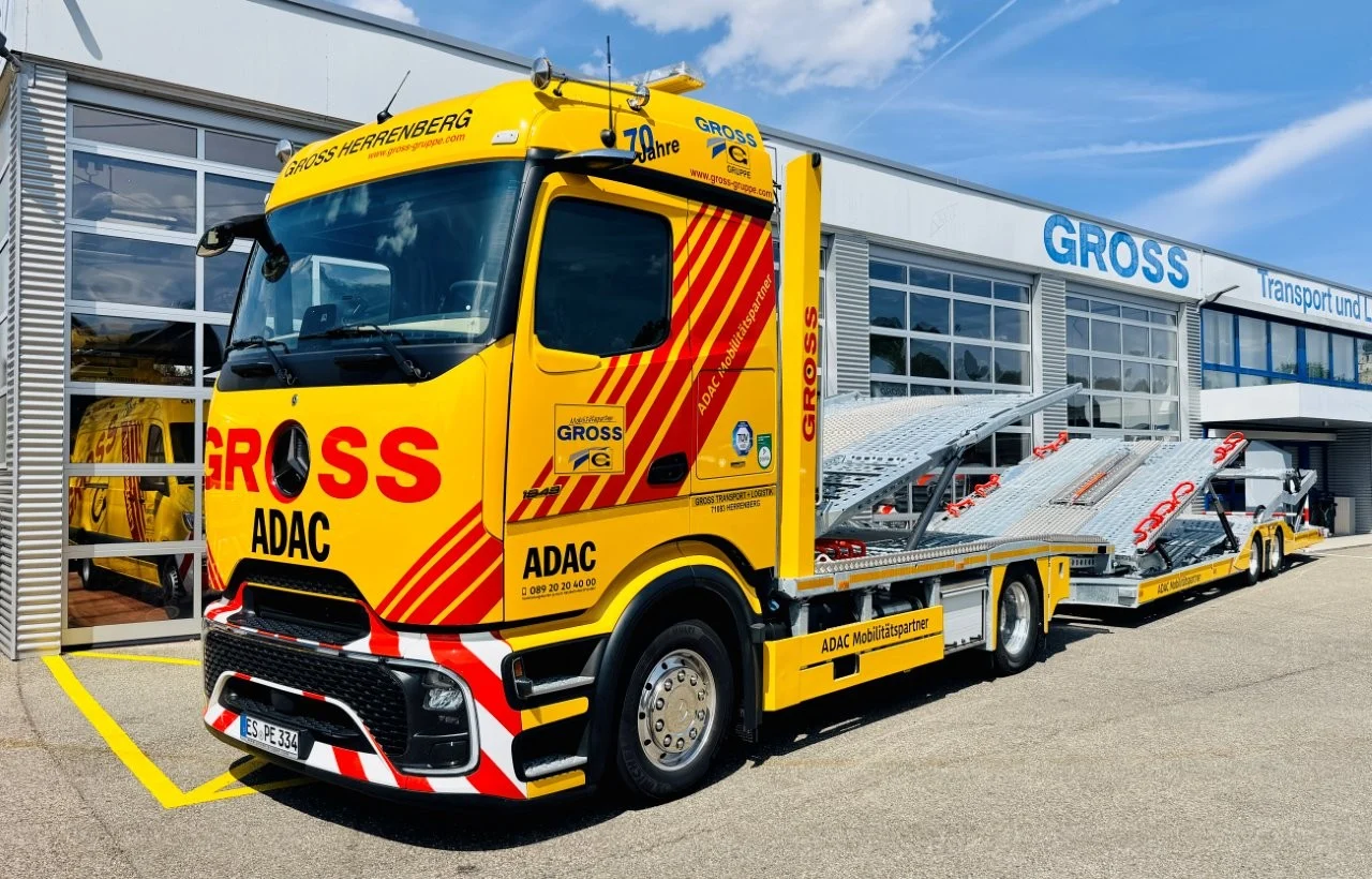 Gelber ADAC Abschleppwagen mit geripptem Anhänger vor einem Gebäude mit der Aufschrift „GROSS Transport und Logistik“.