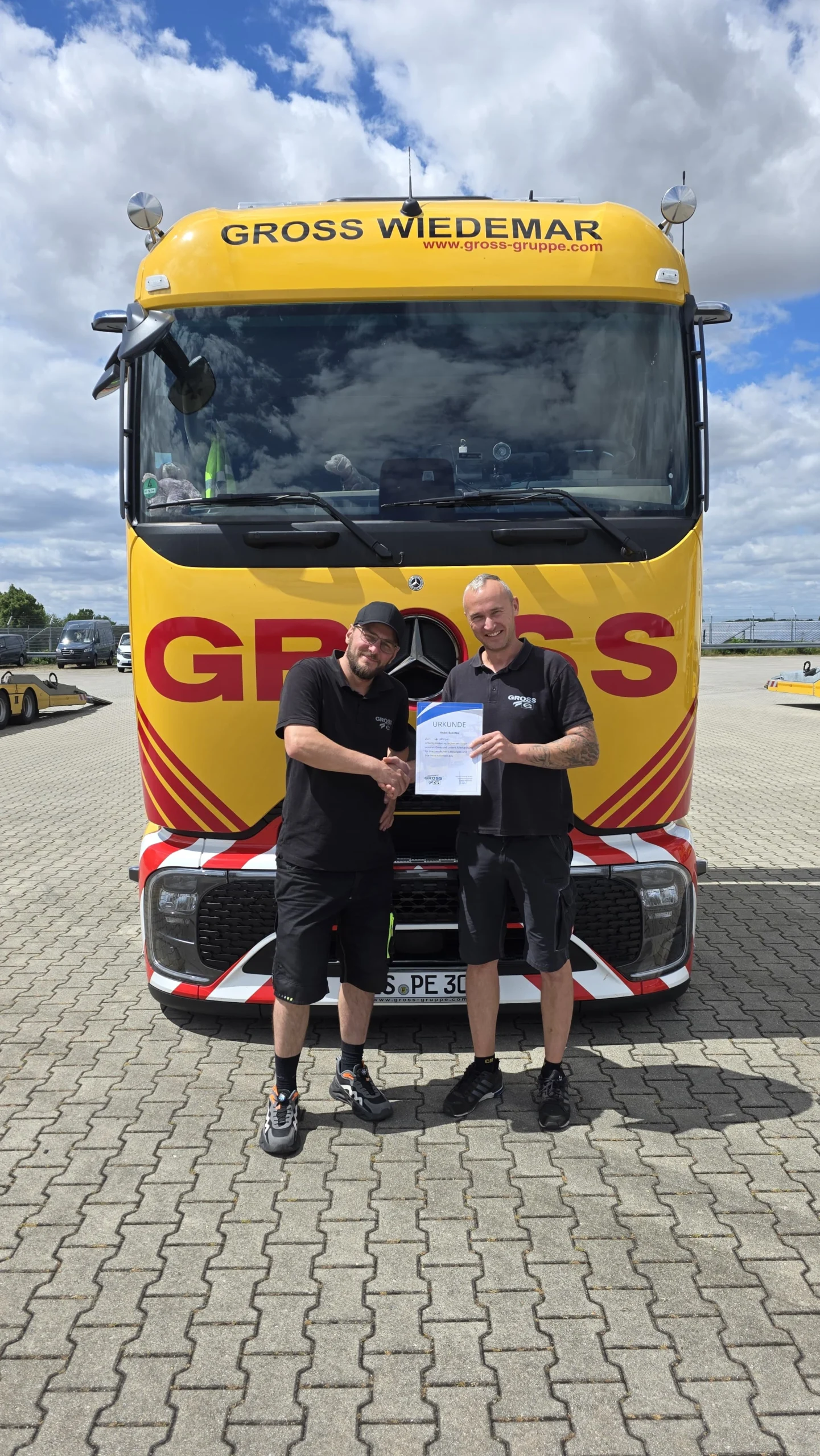 Zwei Männer stehen vor einem gelben Lkw mit dem Schriftzug GROSS WIEDEMAR und halten eine Urkunde.