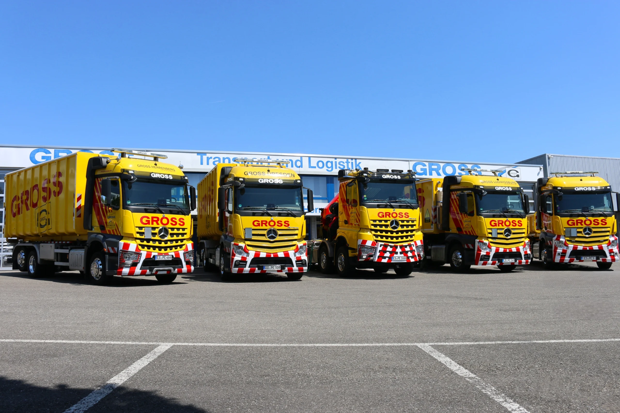 Fünf gelbe Lkw der Firma GRÖSS stehen auf einem Parkplatz vor einem Logistikgebäude unter blauem Himmel.