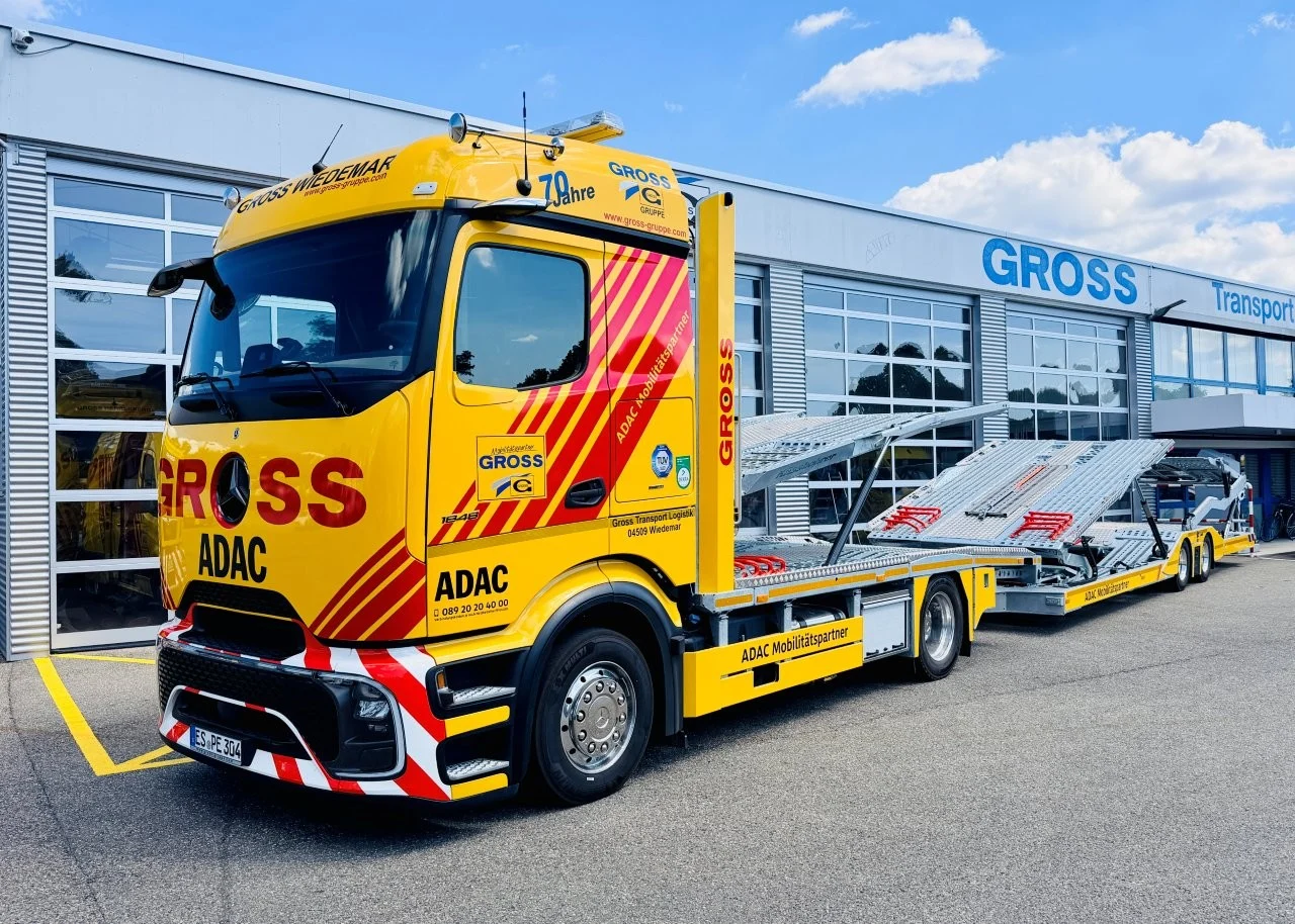 Gelber ADAC-Abschleppwagen mit Ladefläche vor einem modernen Transportgebäude, umgeben von blauem Himmel.