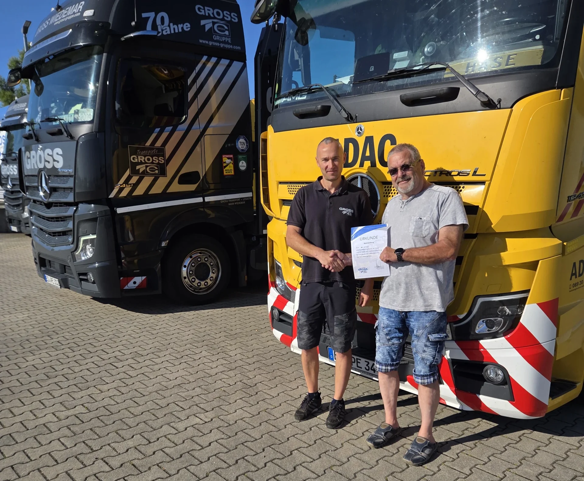 Zwei Männer schütteln sich die Hände vor zwei Lkw, einer in Schwarz und einer in Gelb, mit einem Zertifikat in der Hand.
