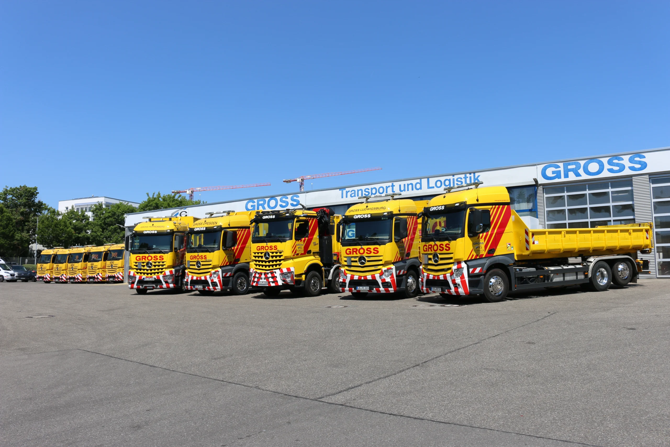 Flotte gelber Lkw der Firma GROSS vor dem Firmengebäude mit dem Schriftzug „Transport und Logistik“ im Hintergrund.