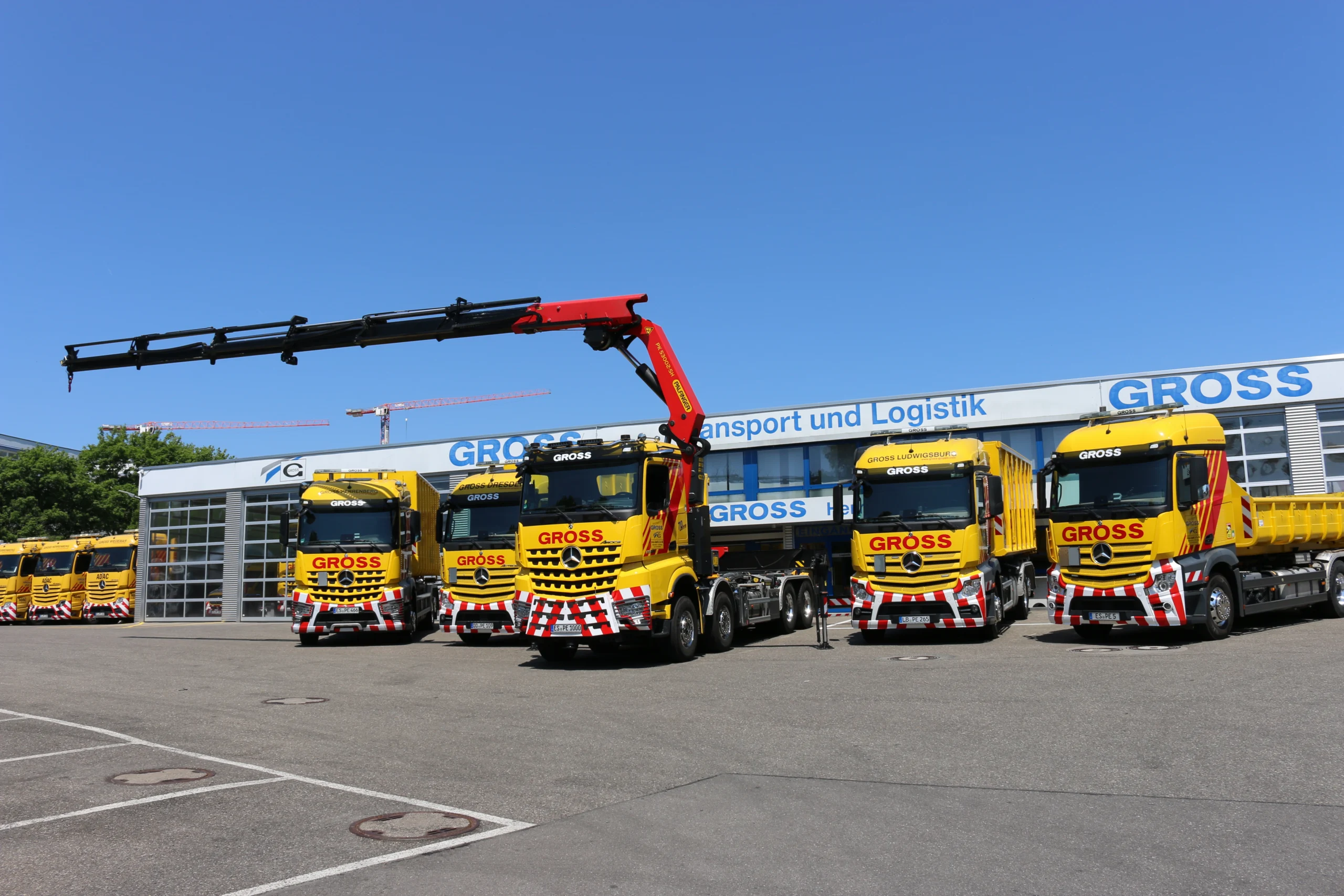 Parkplatz mit gelben Lkw der Firma GROSS, darunter ein Kran, vor dem Bürogebäude im Freien bei blauem Himmel.