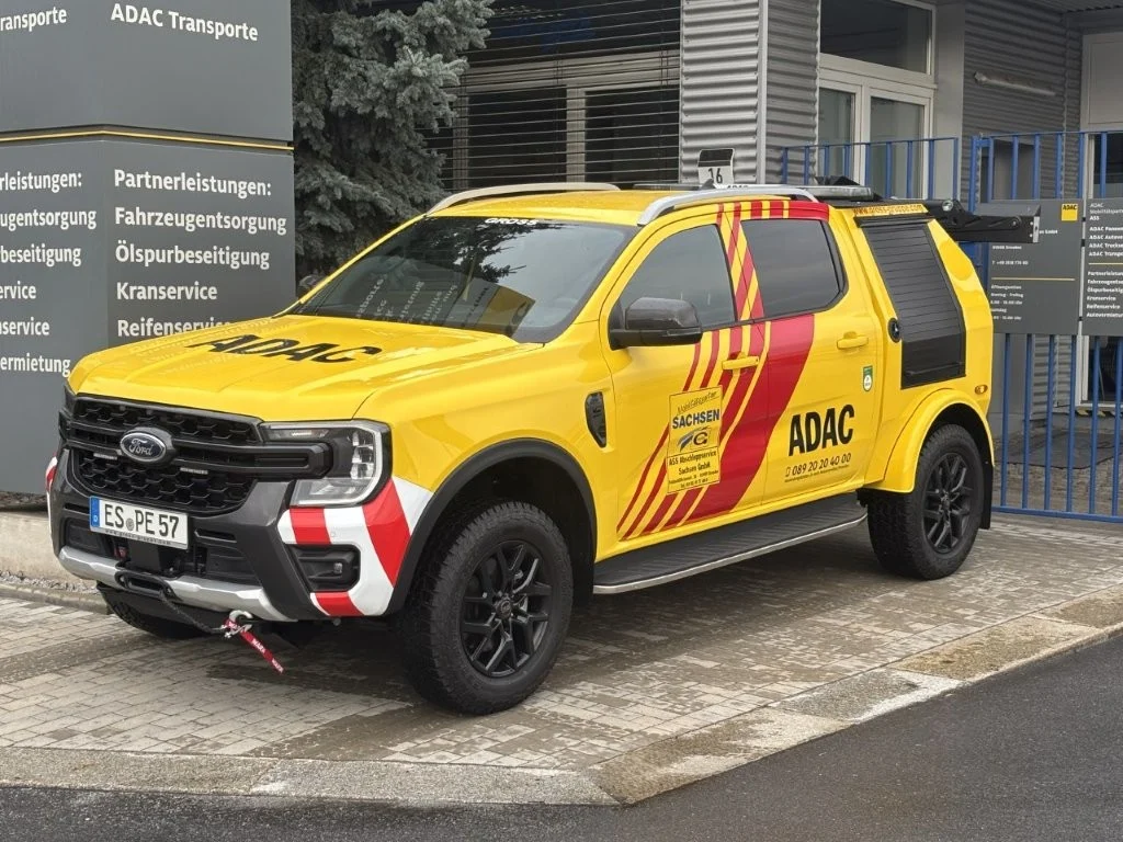 Gelber ADAC-Transporter mit schwarzer Felge vor einer Dienststelle, beschriftet mit ADAC-Logo und Kontaktdaten.