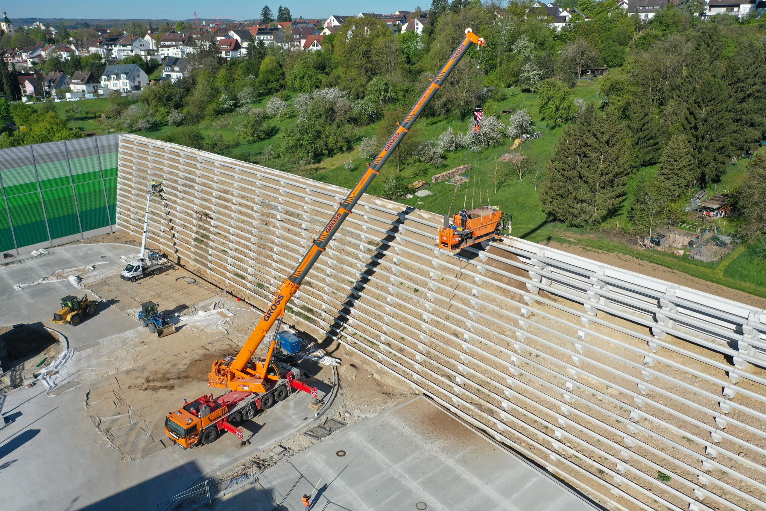 Baustelle mit Kran, der eine orangefarbene Plattform an eine hohe Wand hebt; im Hintergrund grüne Wiesen und Häuser.