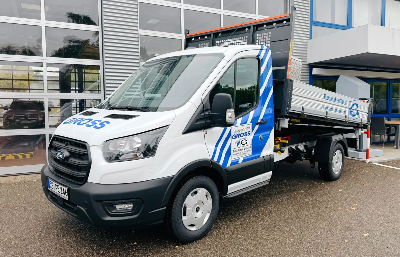 Ein Ford Transporter mit Auflieger in der Firmenfarbe Blau und dem Schriftzug GROSS vor einem modernen Gebäude.